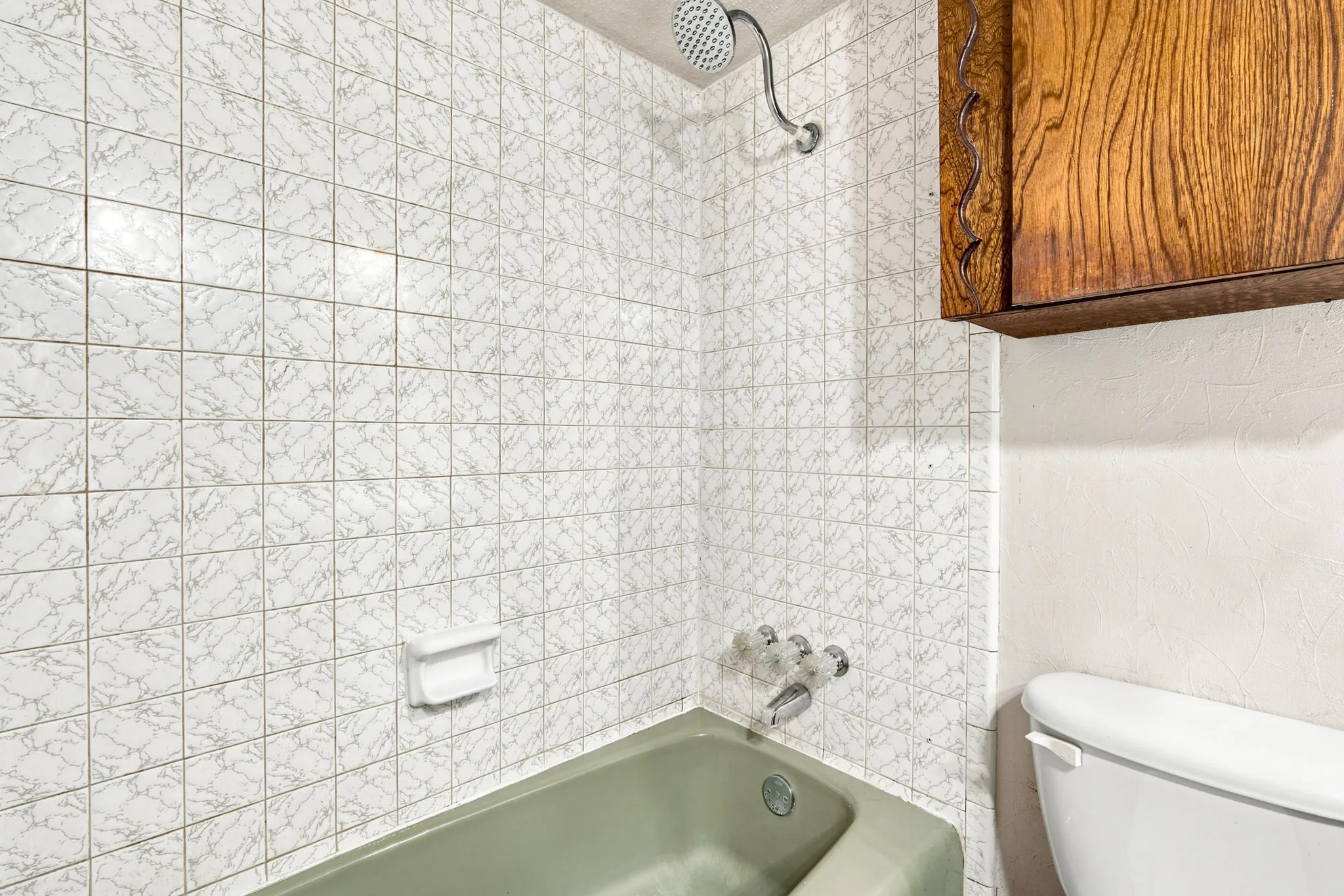 Bathroom featuring a green bathtub with integrated shower, white tiled walls, a chrome showerhead, and a wood-finish wall cabinet
