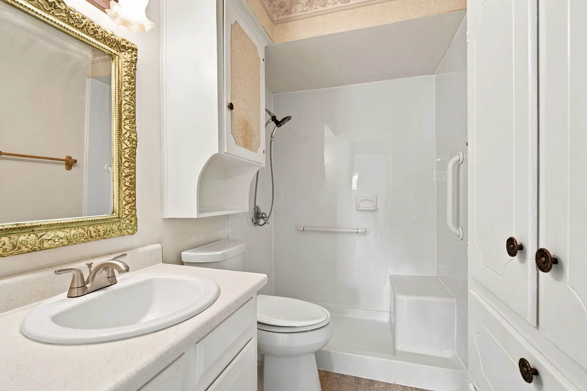 Bathroom featuring a single vanity with a brushed nickel faucet, an ornate gold-framed mirror, an integrated shower system with a built-in bench, and white cabinetry