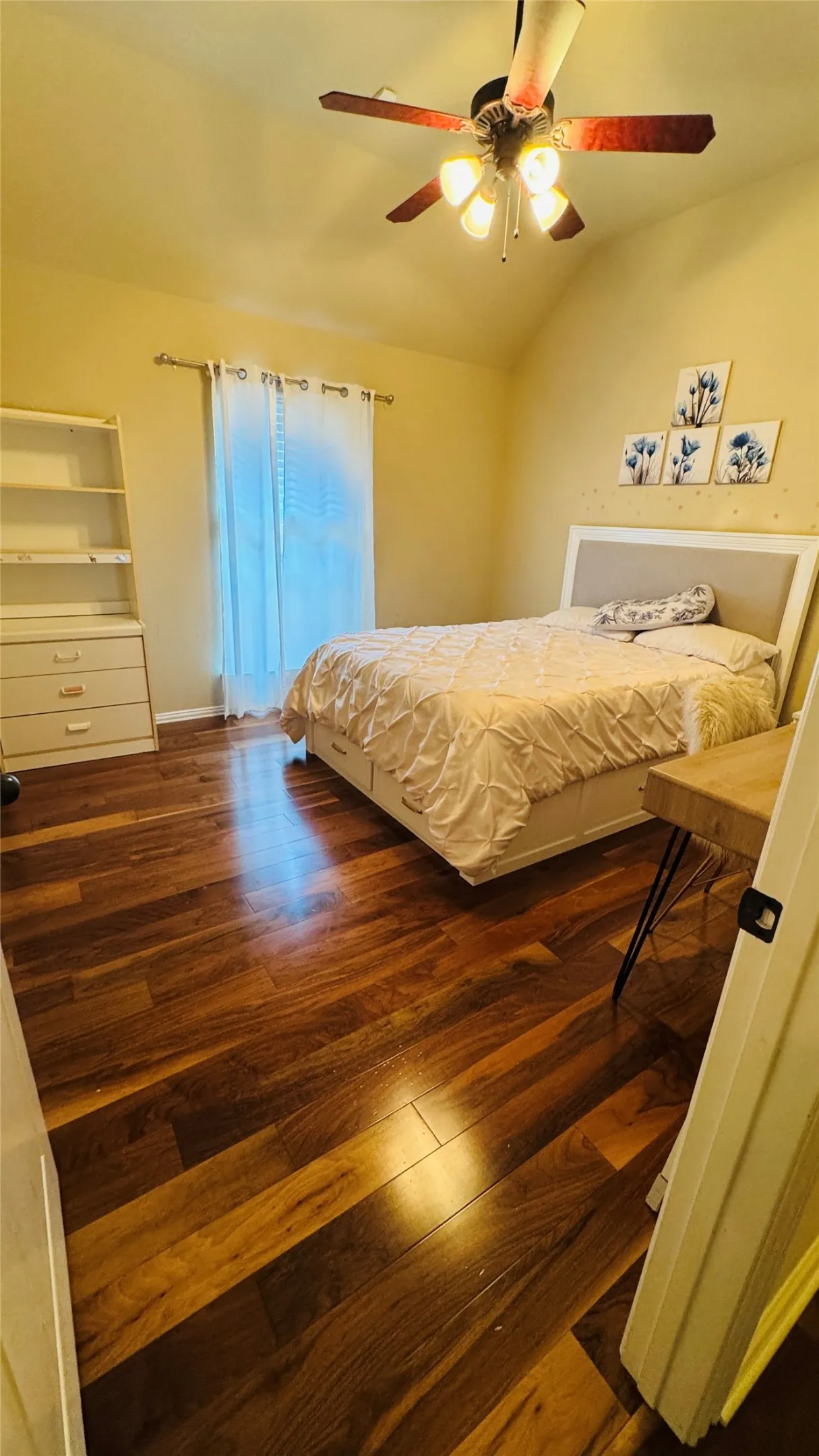 Vaulted ceiling, ceiling fan with integrated lighting, wood-finish flooring, white window treatments, and a built-in shelving unit