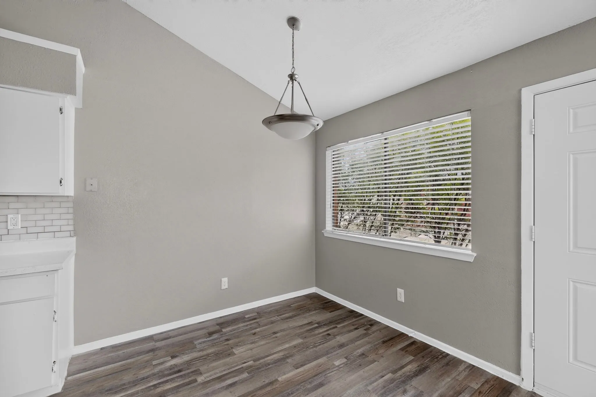 Dining area featuring wood-finish flooring, a large window with blinds, a pendant light fixture, and white cabinetry with a subway tile backsplash