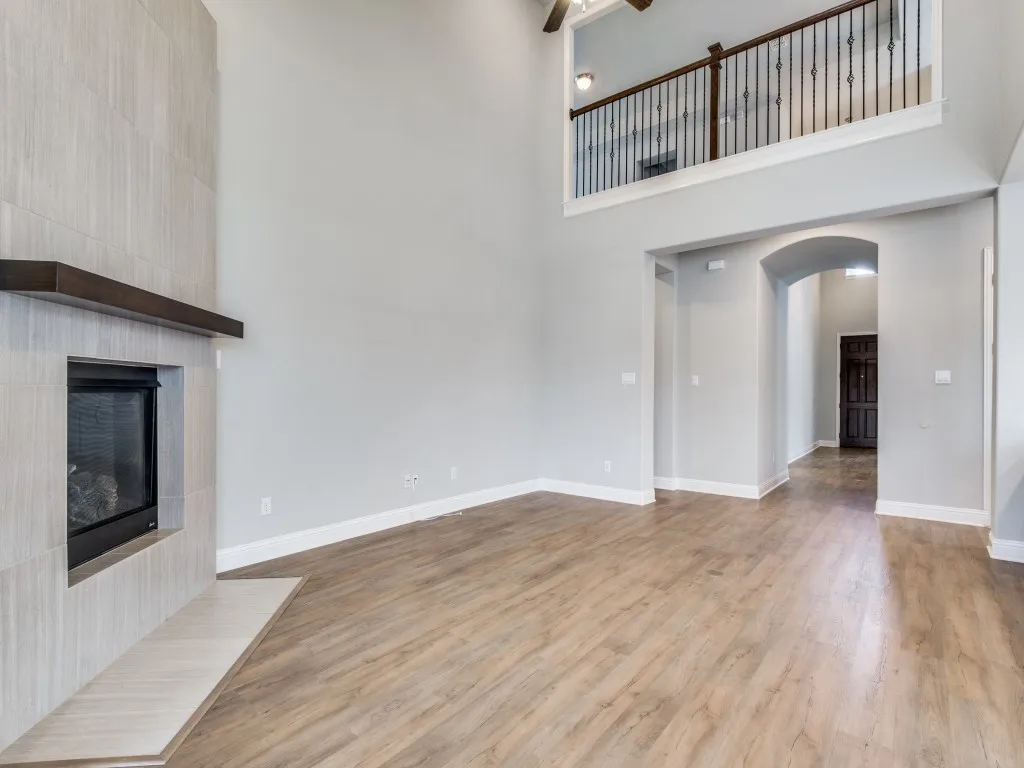 Living area featuring high ceilings, wood-finish flooring, a contemporary fireplace with a light-toned tile surround and dark wood mantle, an arched entryway, and an upper-level overlook with wrought iron balusters