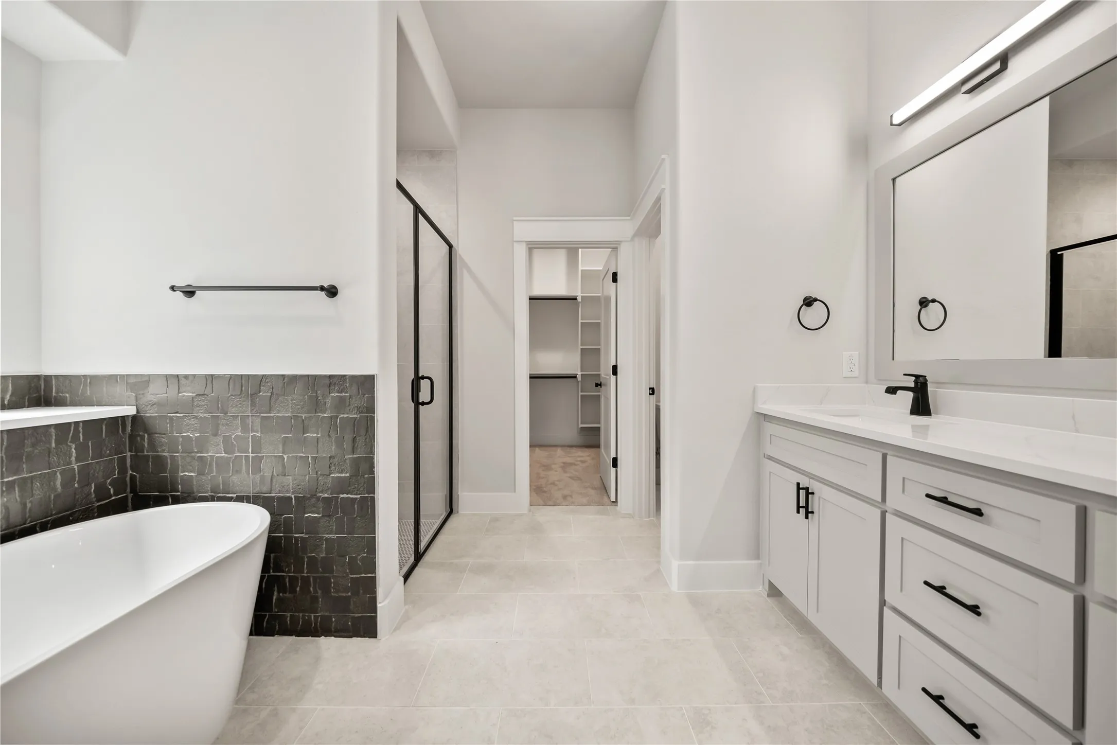 Bathroom featuring a freestanding tub, dark tile wainscoting, glass-enclosed shower, single vanity with quartz countertop, and light tile flooring