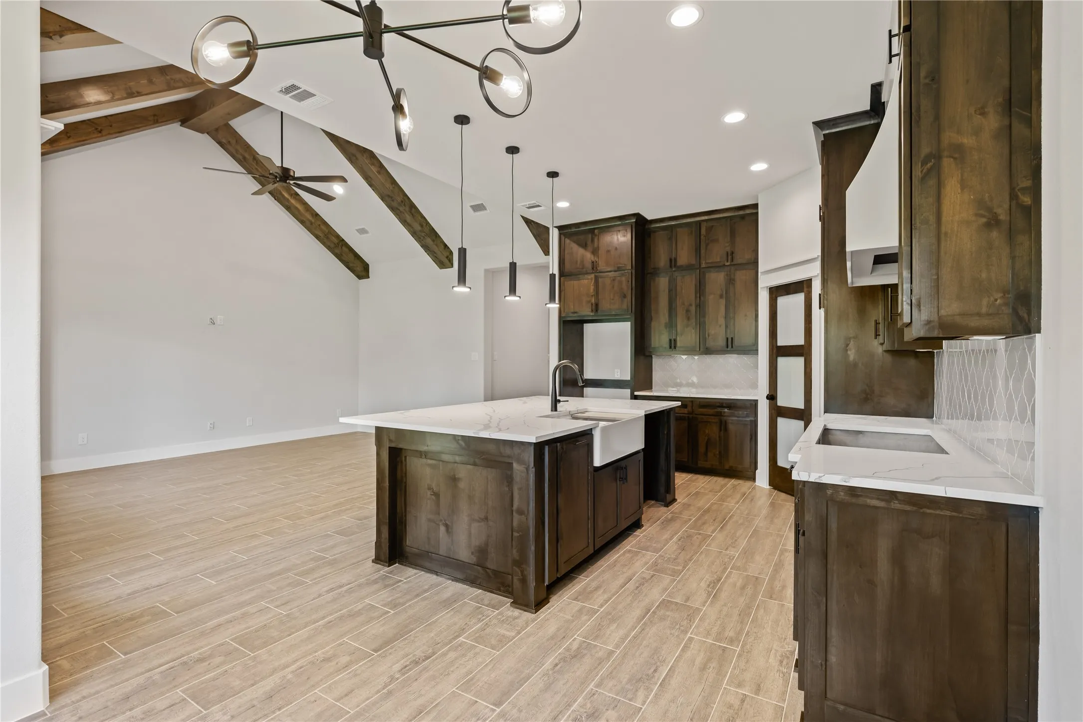 Open-concept kitchen and living area featuring vaulted ceilings with exposed wood beams, a contemporary chandelier, recessed lighting, a kitchen island with an undermount farmhouse sink, and a cooktop with a range hood