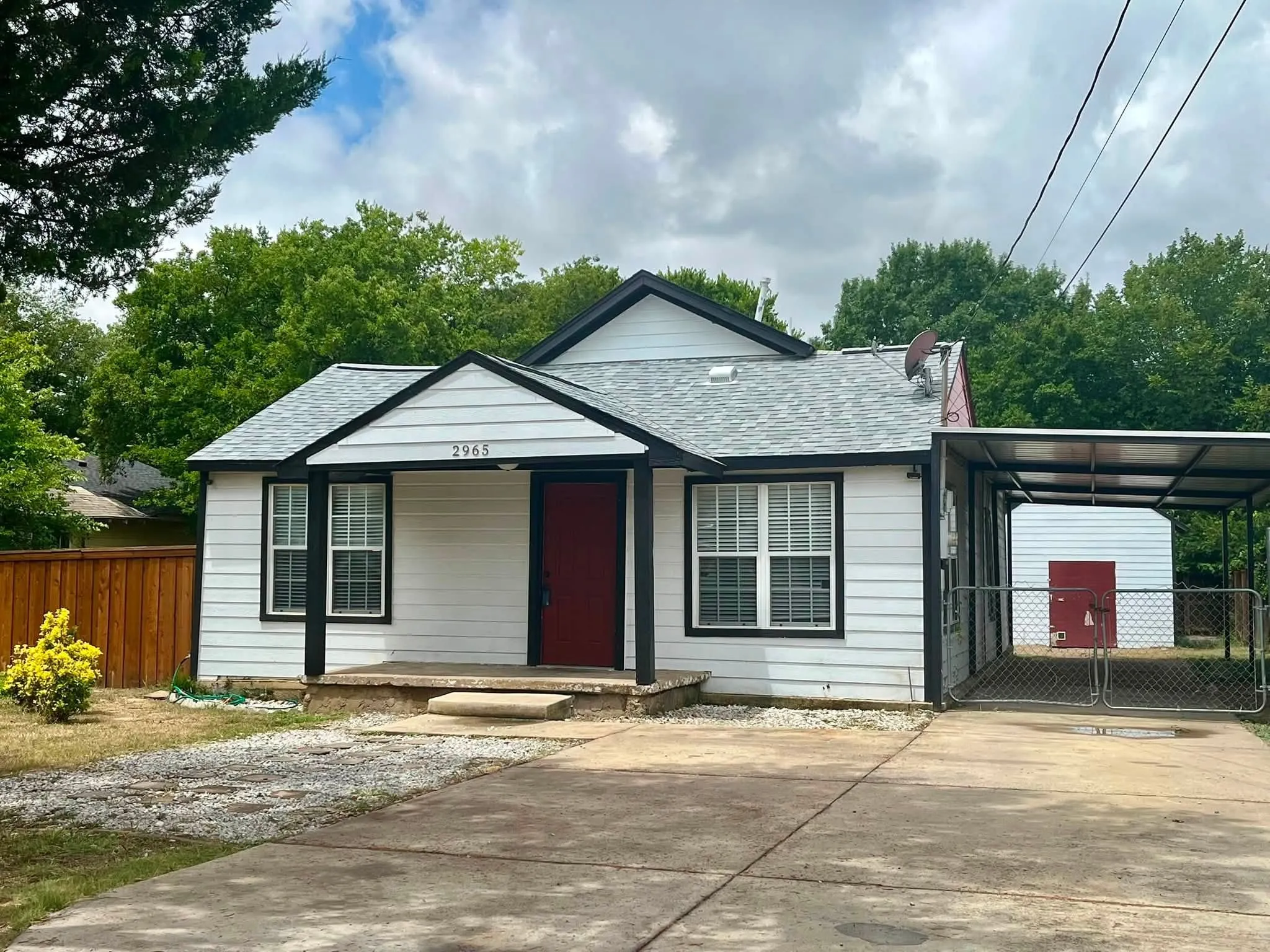 White exterior siding with black trim, a red entry door, a covered carport, and a detached shed