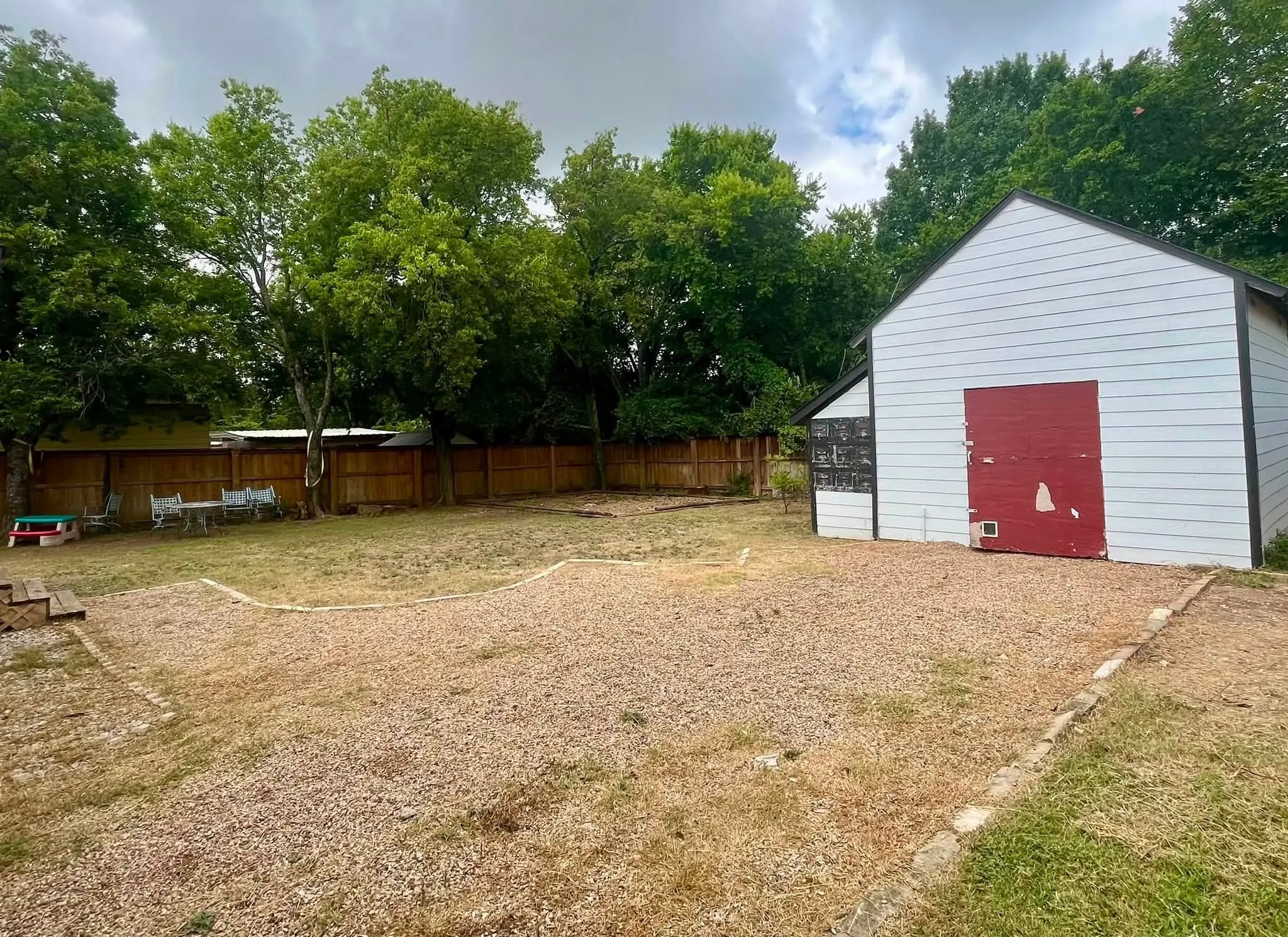 White-sided outbuilding with red doors, gravel pad, and partially fenced yard featuring mature trees