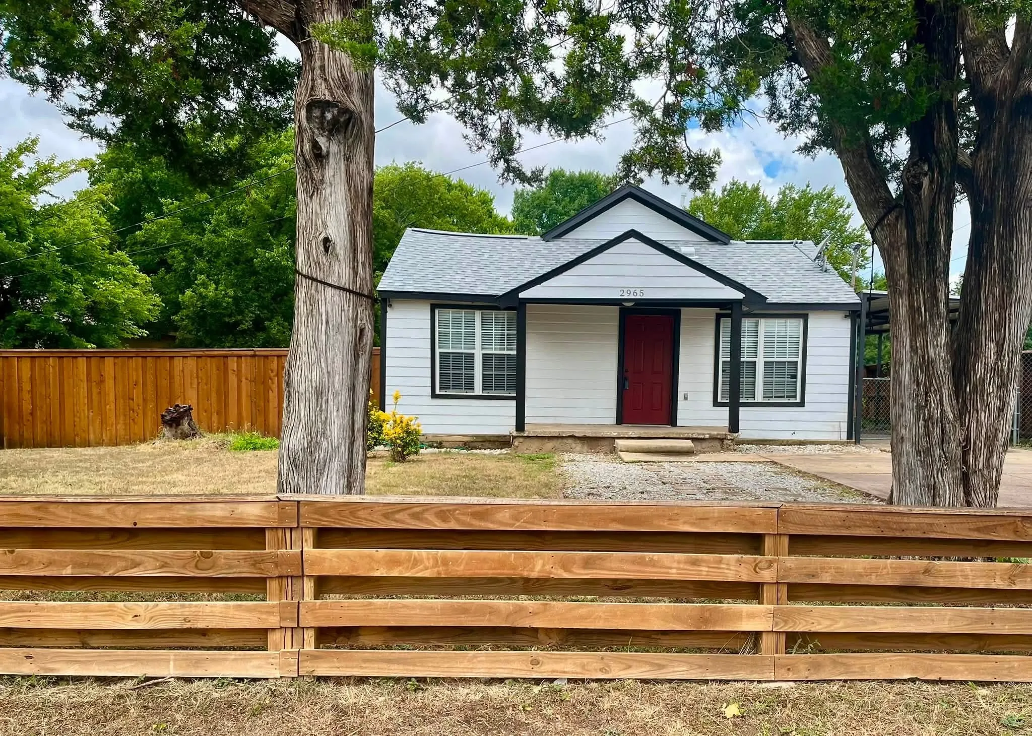 White siding residence with a red entry door, framed by mature trees and a horizontal wood fence