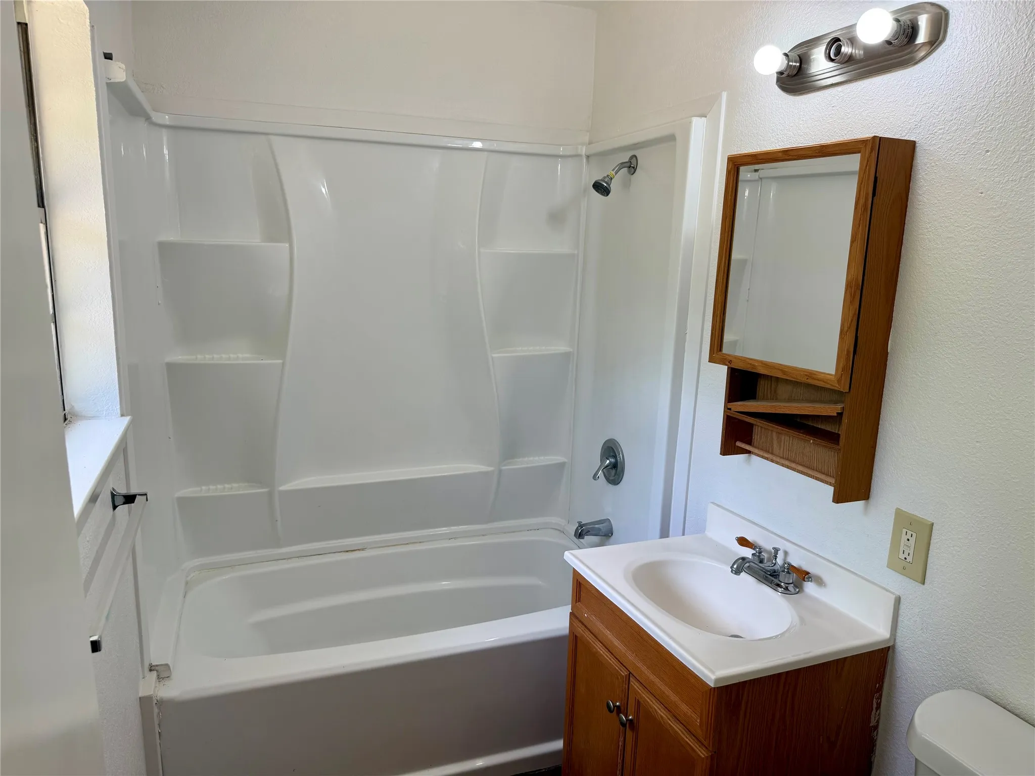 Bathroom featuring a shower-tub combination with integrated shelving, wood-finish vanity with countertop and sink, and a wood-framed medicine cabinet