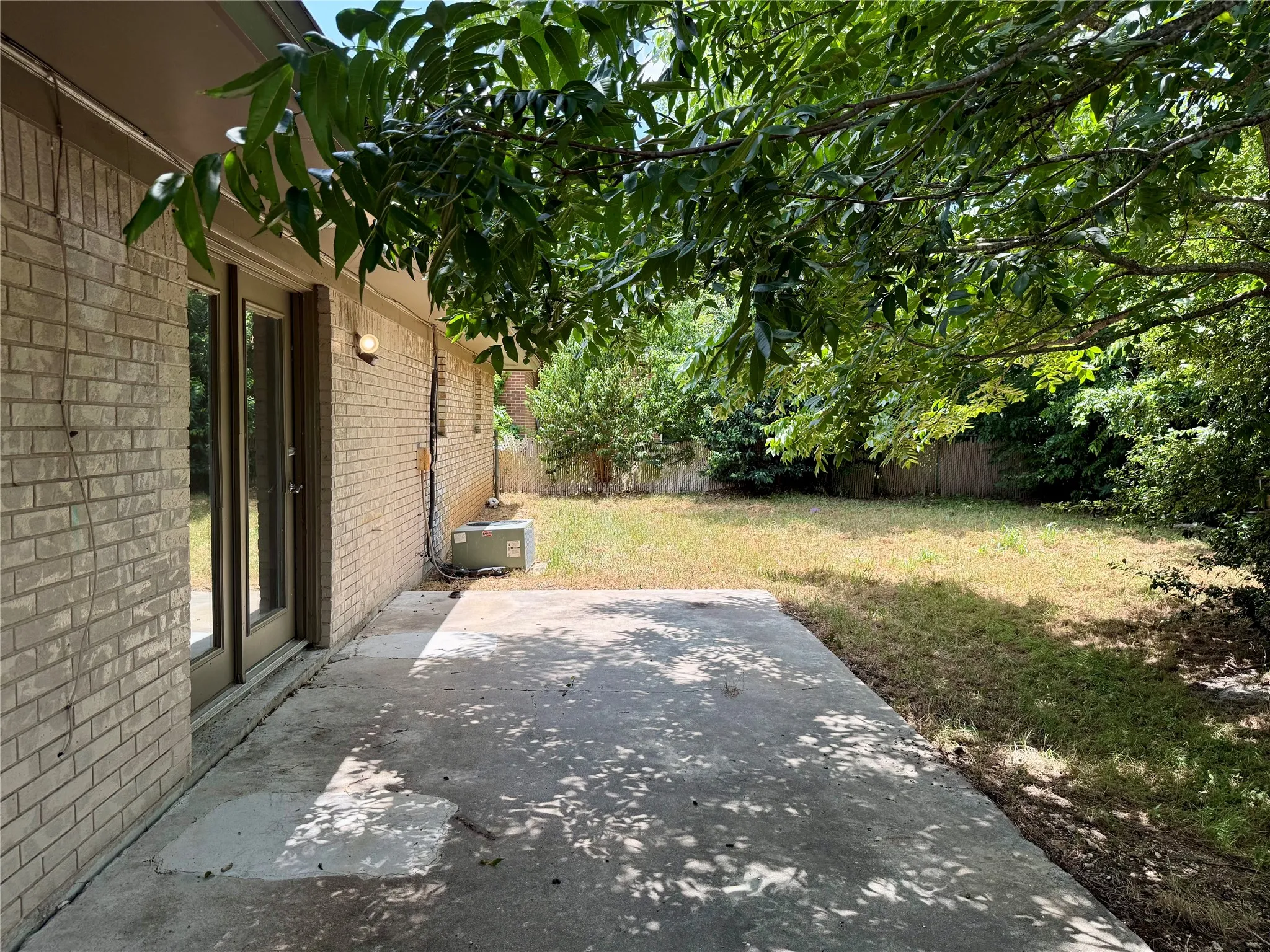 Concrete patio area with brick exterior and dual glass-paneled doors, surrounded by lush mature trees and a grassy yard