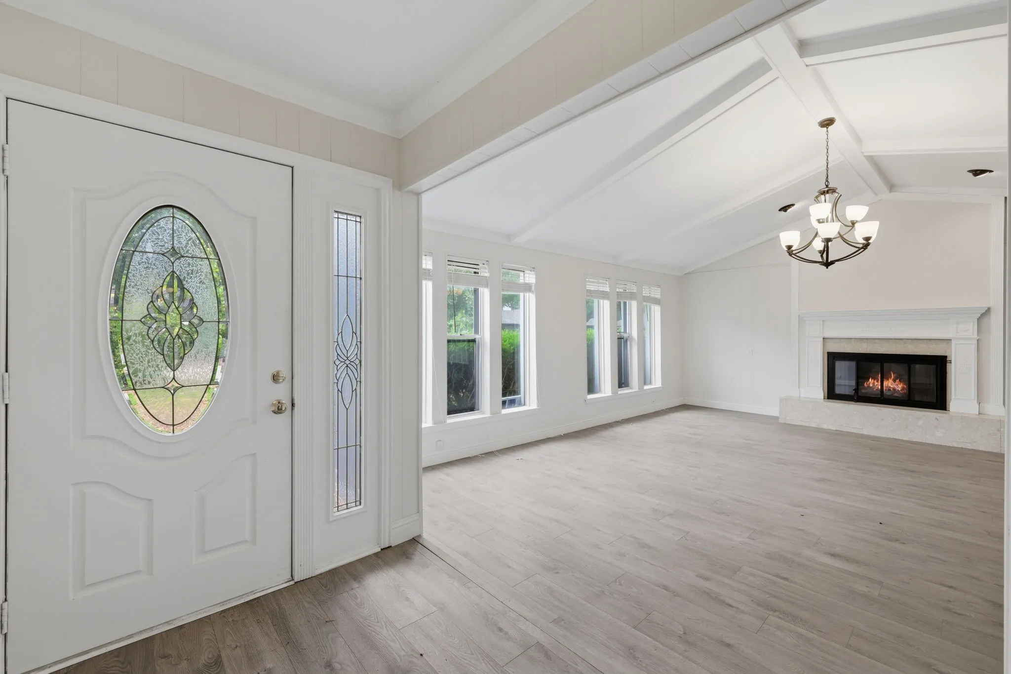 Entryway featuring a decorative glass door with a matching sidelight, opening into a spacious room with vaulted ceilings, wood-finish flooring, a fireplace with a white mantel, and multiple windows along the wall