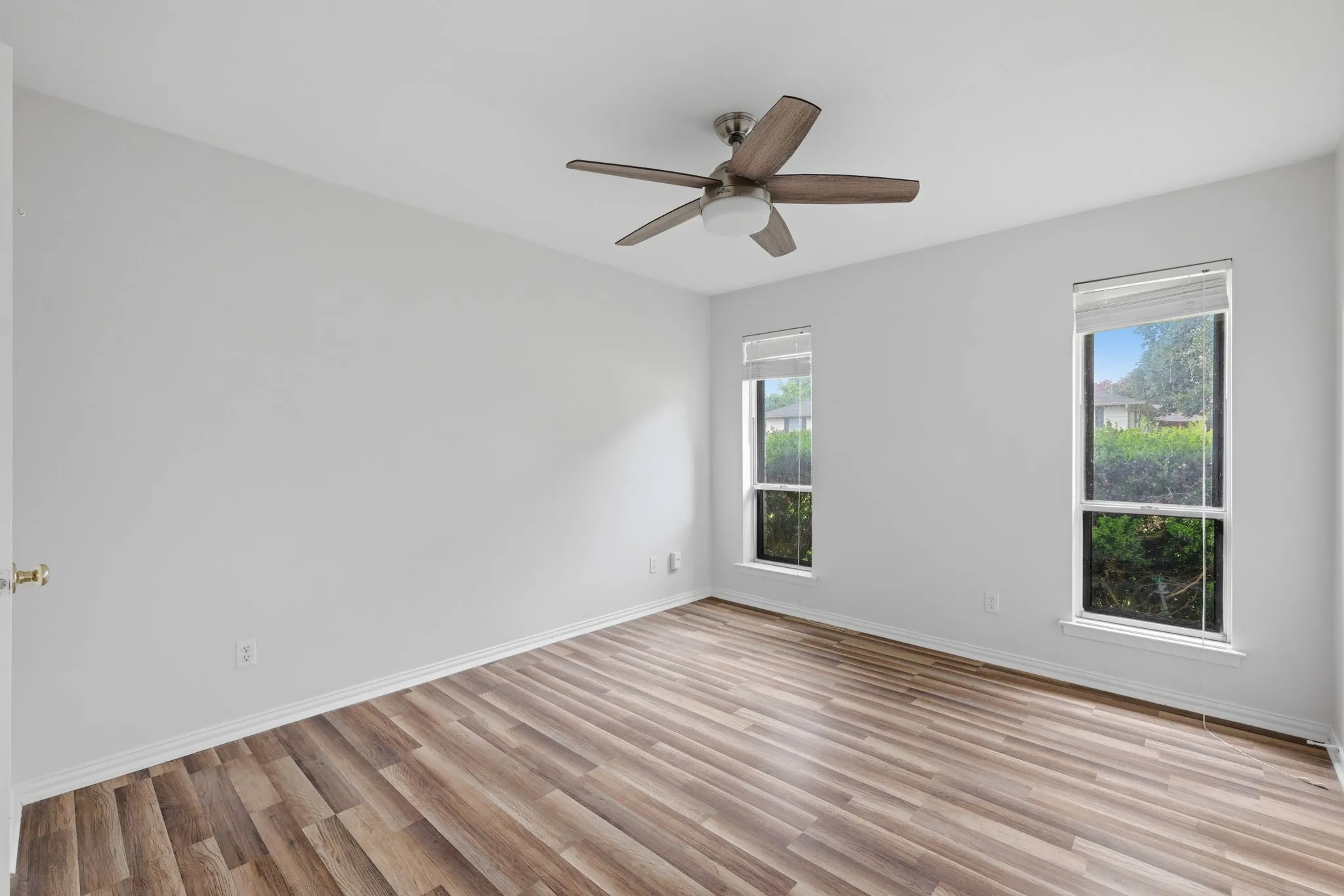 Spacious room featuring wood-finish flooring, two windows with blinds, a ceiling fan with integrated lighting, and white baseboards