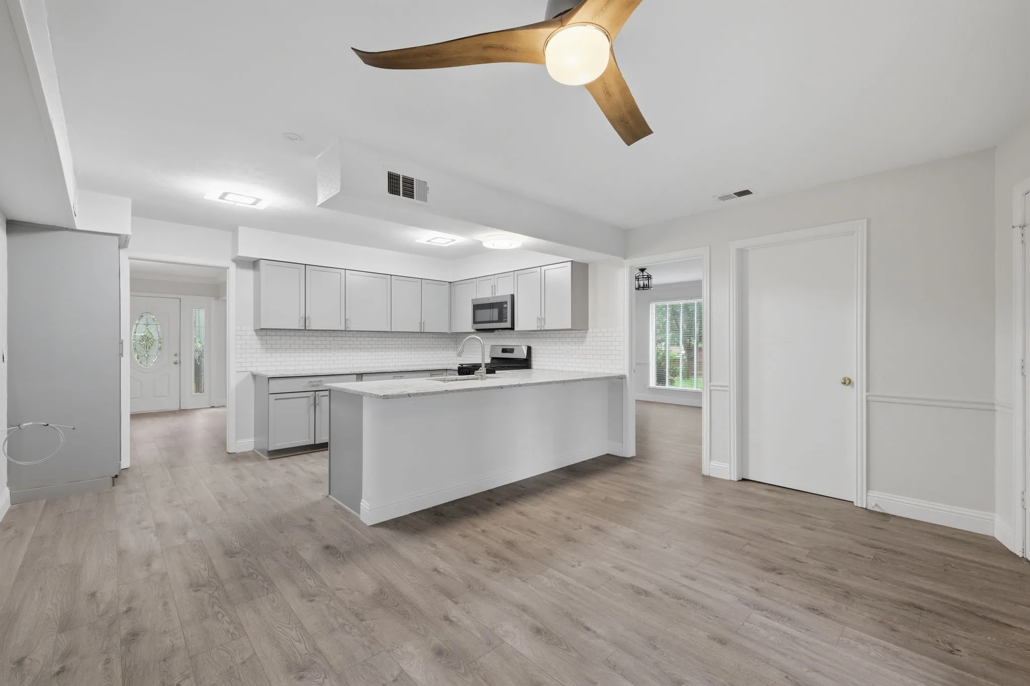 Open-concept kitchen featuring gray cabinetry, a central island with a sink, stainless steel microwave, white subway tile backsplash, and light wood-finish flooring