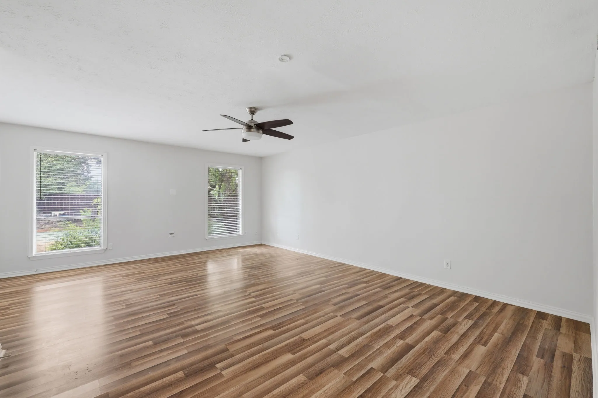 Spacious room featuring wood-finish flooring, two large windows with horizontal blinds, a ceiling fan with integrated lighting, and recessed overhead lighting