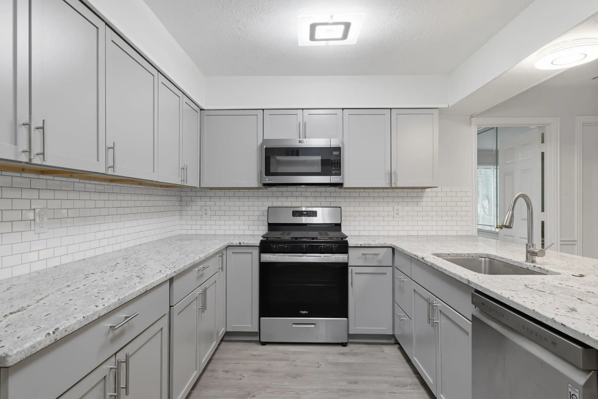 Kitchen featuring light gray shaker cabinetry, white subway tile backsplash, light stone-finish countertops, stainless steel appliances, and wood-finish flooring