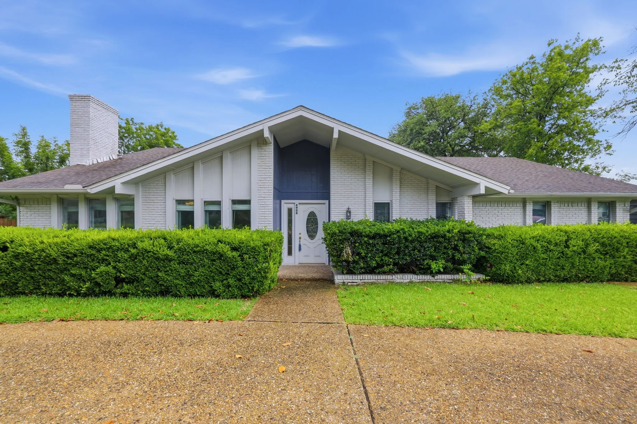 Mid-century modern exterior featuring white painted brick, an A-frame portico with blue siding, a white front door with an oval glass insert, a prominent brick chimney, and a gravel-finish driveway