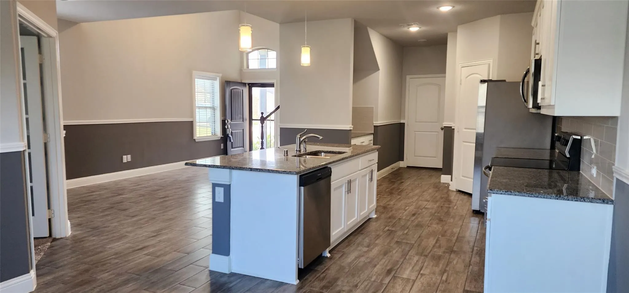 Spacious interior featuring wood-finish flooring throughout, a kitchen island with stone countertop and integrated sink, white cabinetry with silver-tone hardware, stainless steel appliances, and a two-tone wall treatment with wainscoting