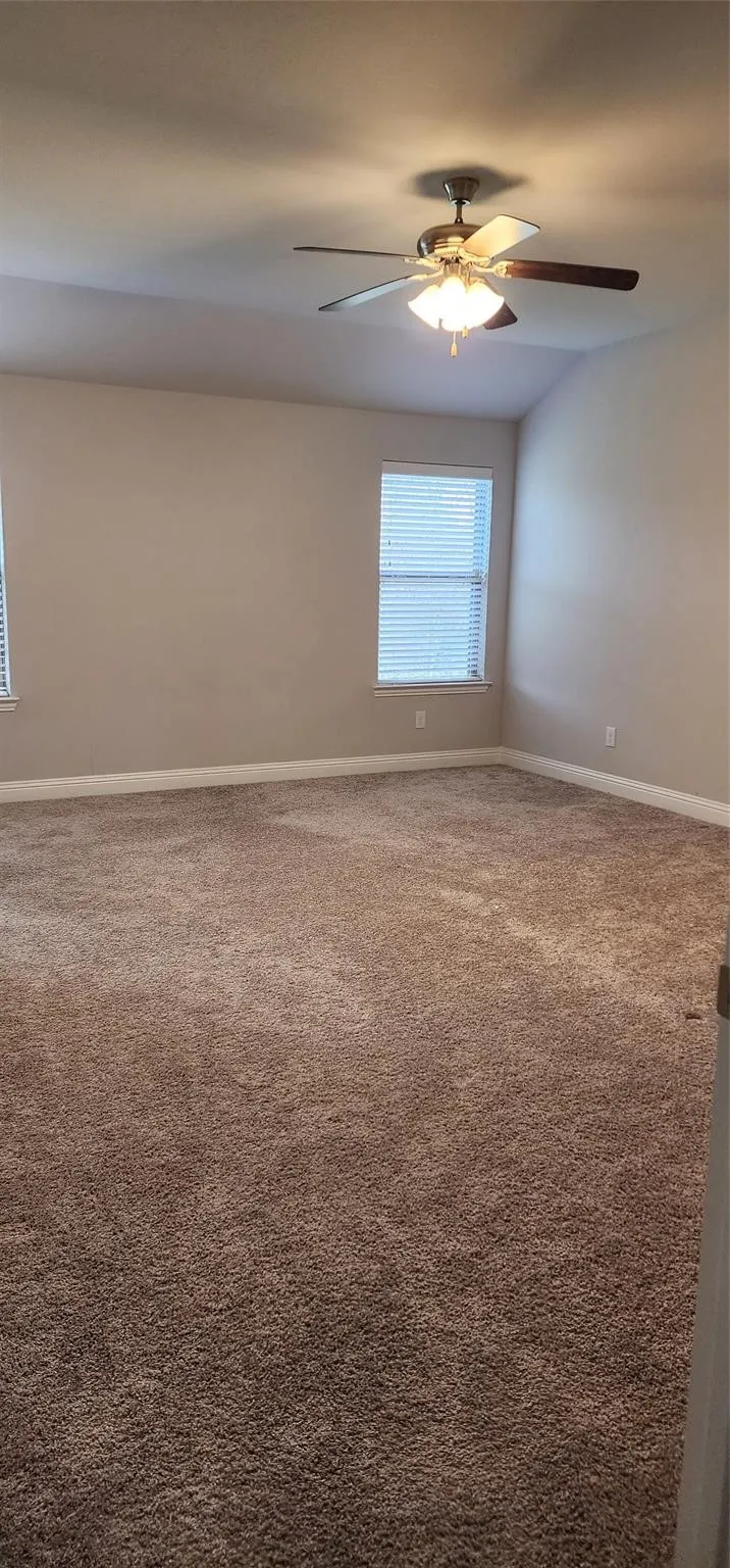 Spacious room featuring neutral carpeting, light-toned walls, two windows with blinds, and a ceiling fan with integrated lighting