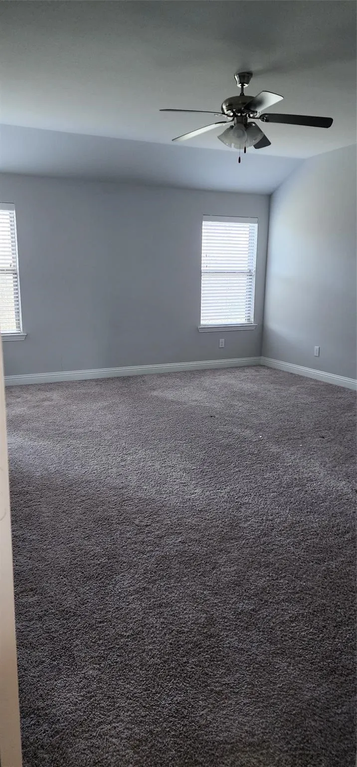 Carpeted interior room featuring a ceiling fan with integrated lighting, two windows with blinds, a vaulted ceiling, and light gray wall paint