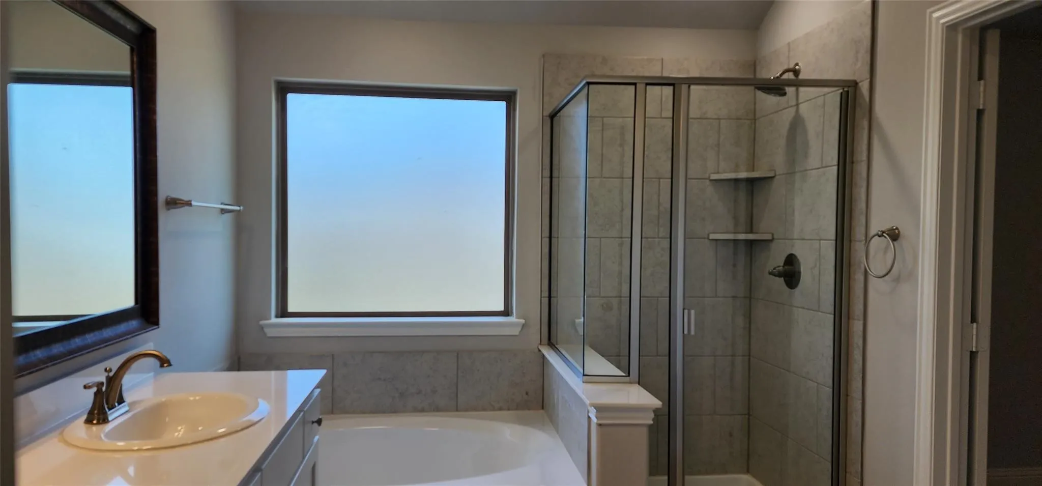 Spacious bathroom featuring a built-in tub, separate glass-enclosed shower with tile surround and integrated shelving, single vanity with oval sink and bronze-finish faucet, and a large mirror