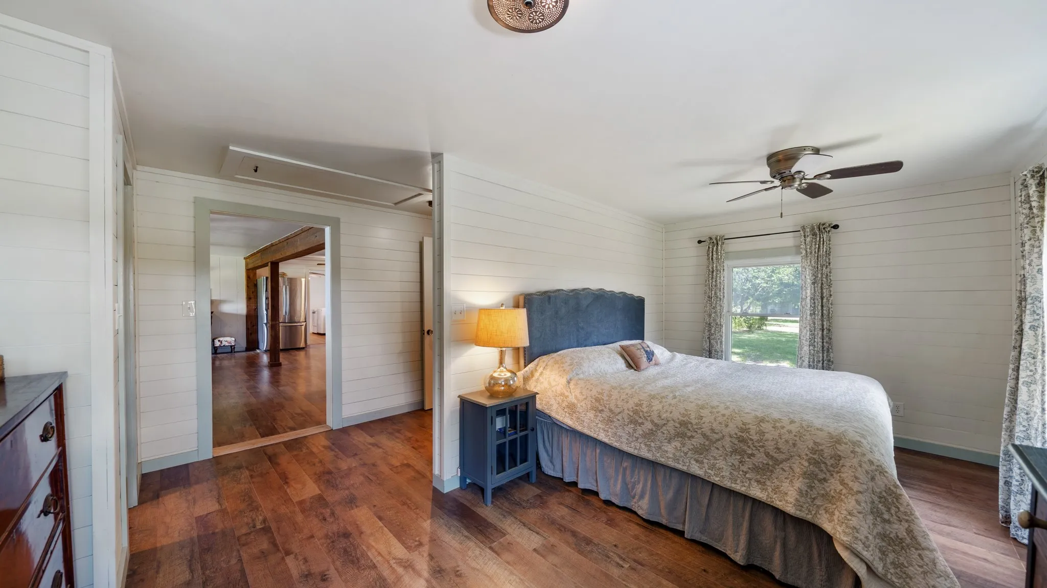 Wood-finish flooring throughout, shiplap walls, ceiling fan, and window with outdoor greenery visible