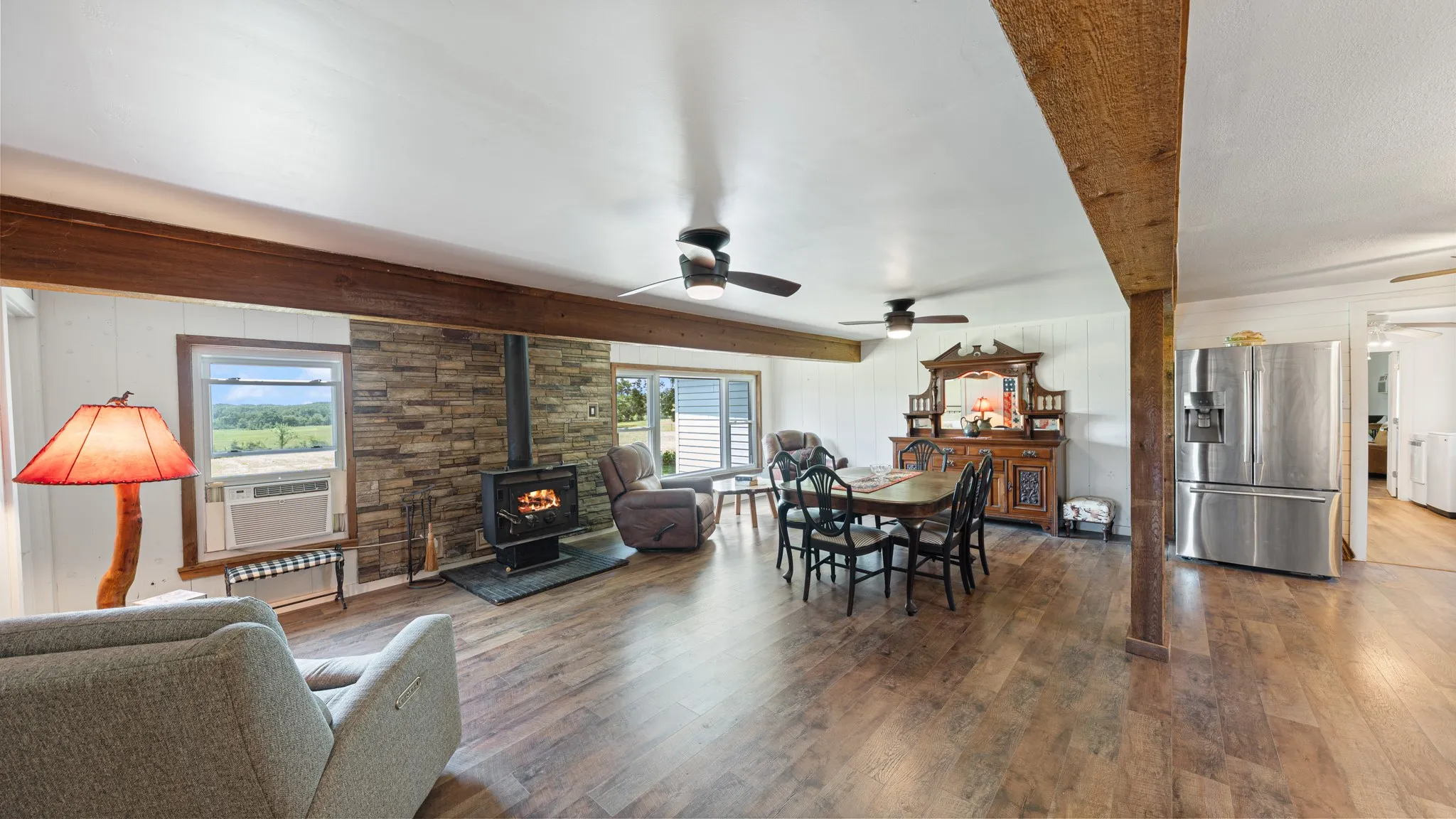 Open-concept interior featuring wood-finish flooring, exposed wood ceiling beams, a stone accent wall, and a built-in wood-burning stove