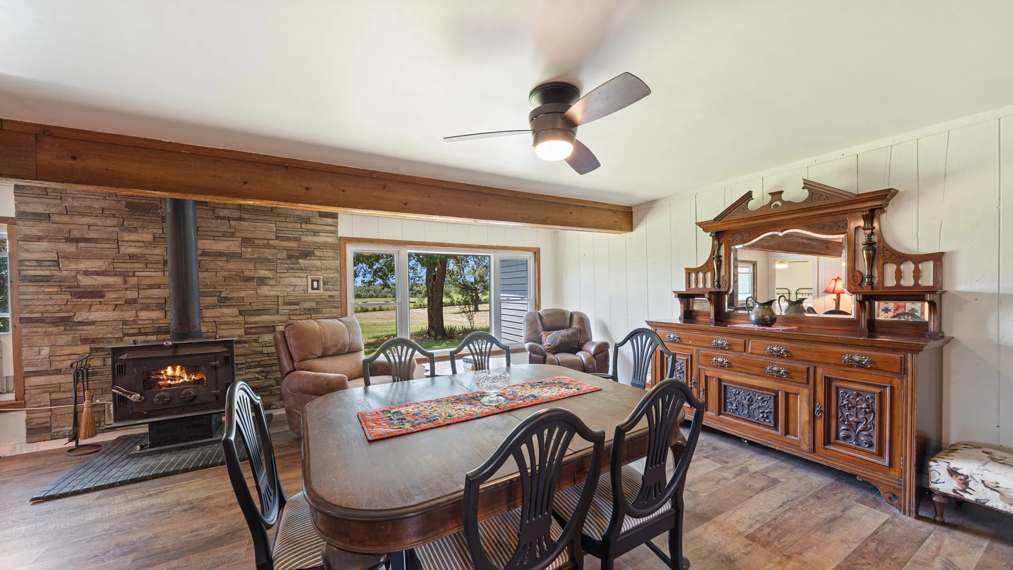 Spacious room featuring a stone-clad wood-burning stove, wood-finish flooring, a wood beam, and panoramic windows