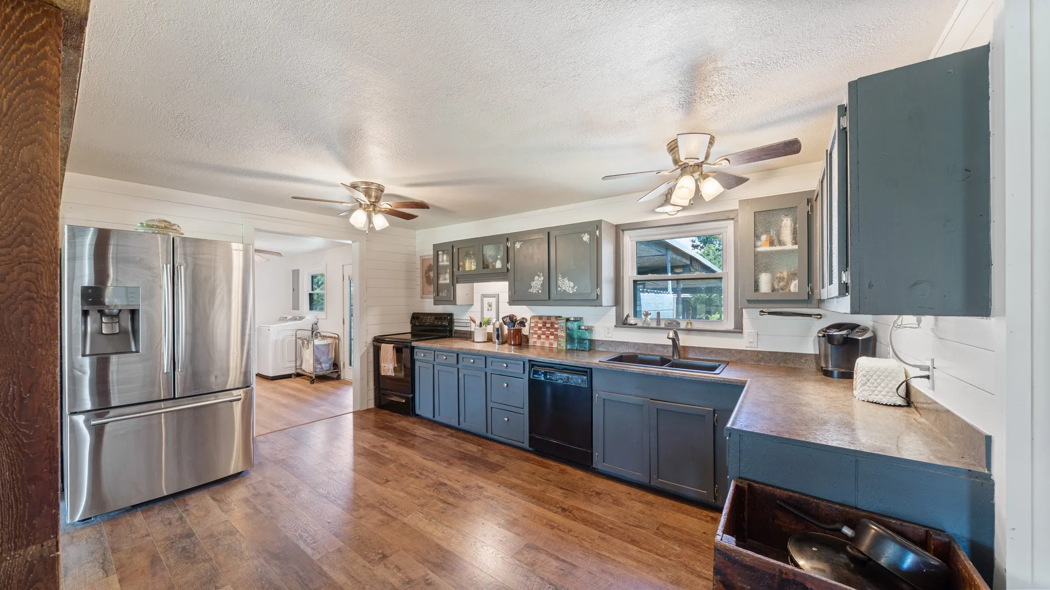 Open-concept kitchen featuring wood-finish flooring, stainless steel refrigerator, dark cabinetry with some glass-front uppers, and ceiling fans with light fixtures