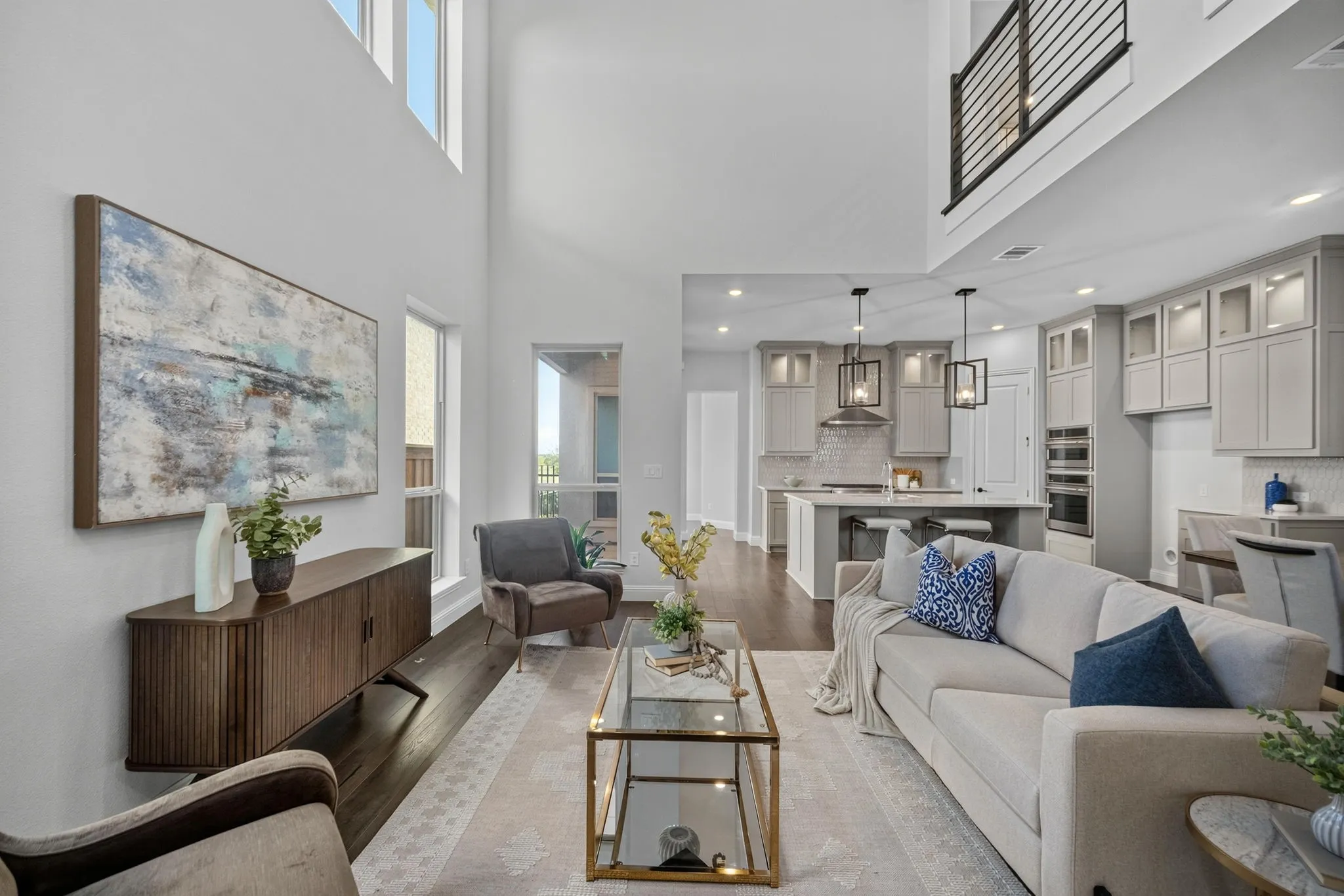 Open-concept living area featuring high ceilings, wood-finish flooring, a modern kitchen with light cabinetry, a kitchen island with seating, and a second-story interior balcony with metal railing