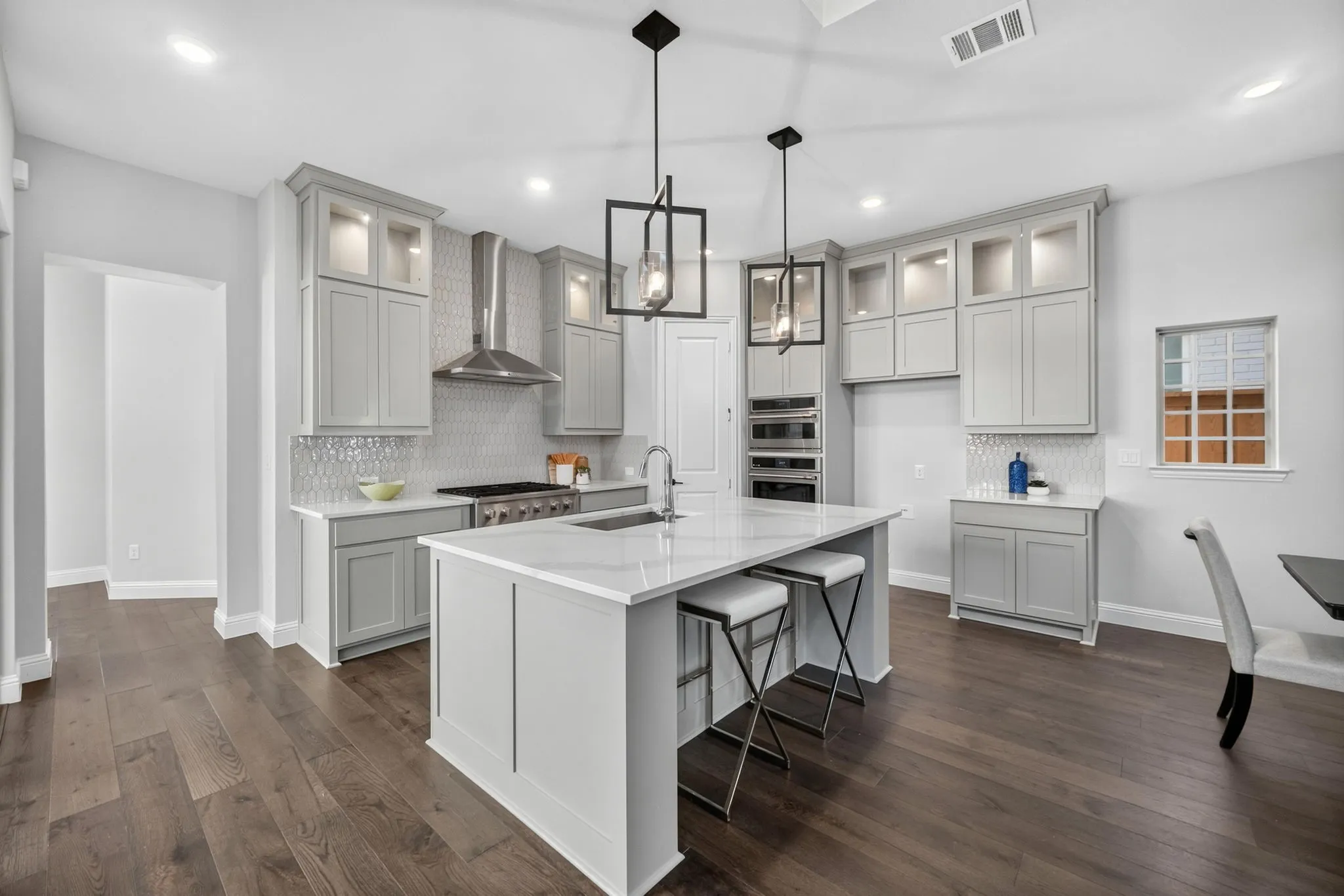 Modern kitchen featuring a large central island with seating, wood-finish flooring, light gray cabinetry with glass accents, and a white tile backsplash
