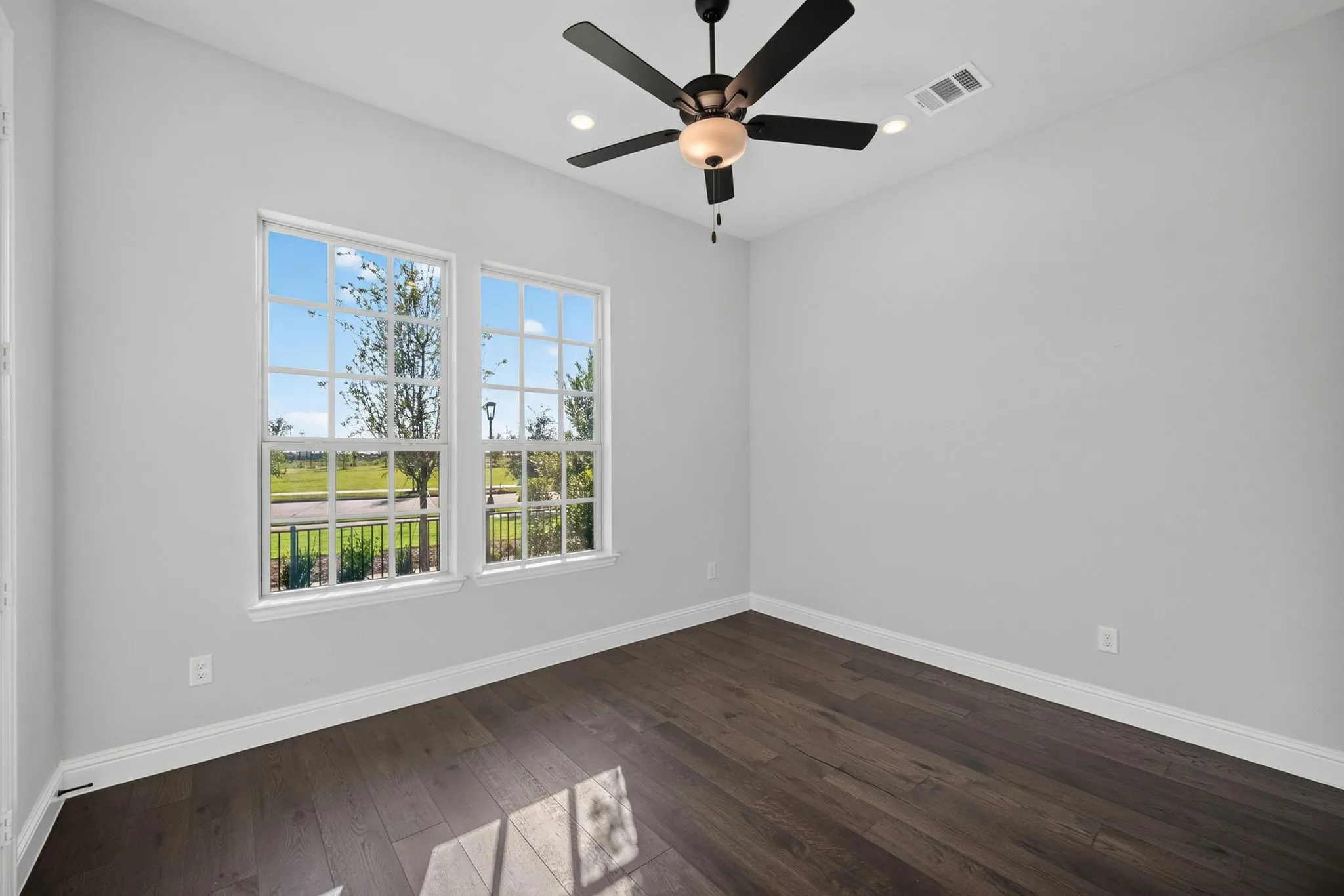 First floor bedroom/study featuring two large windows, beautiful wood-finish flooring, light gray walls, closet and a ceiling fan with integrated lighting