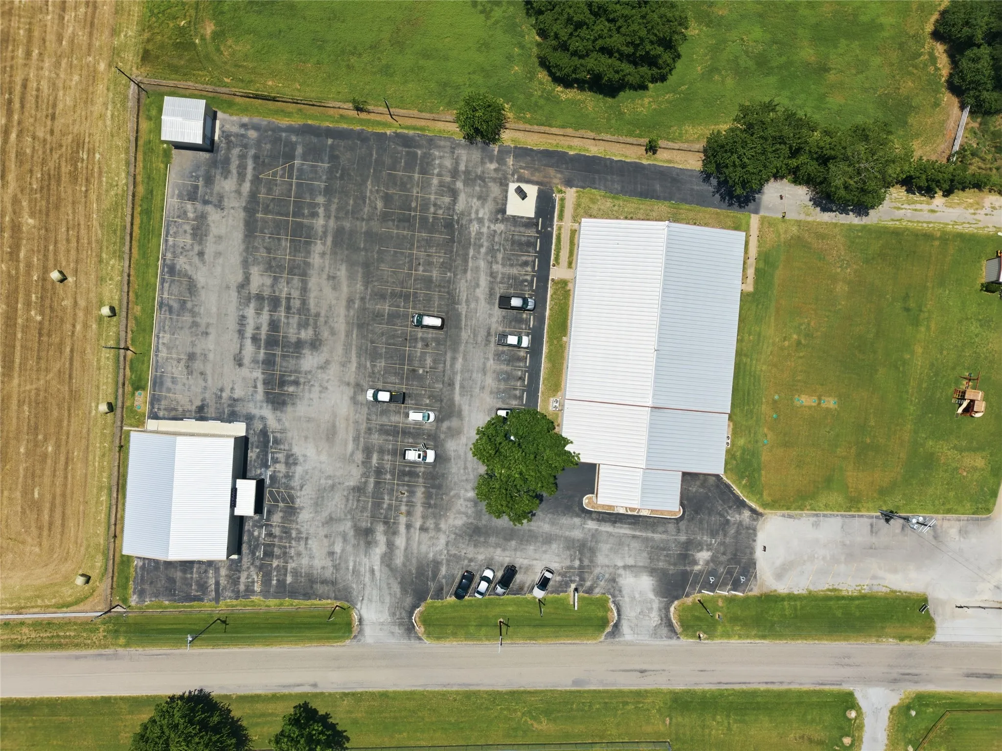 Expansive asphalt parking lot with marked spaces, two large commercial buildings featuring metal roofs, green lawn areas, and direct street access