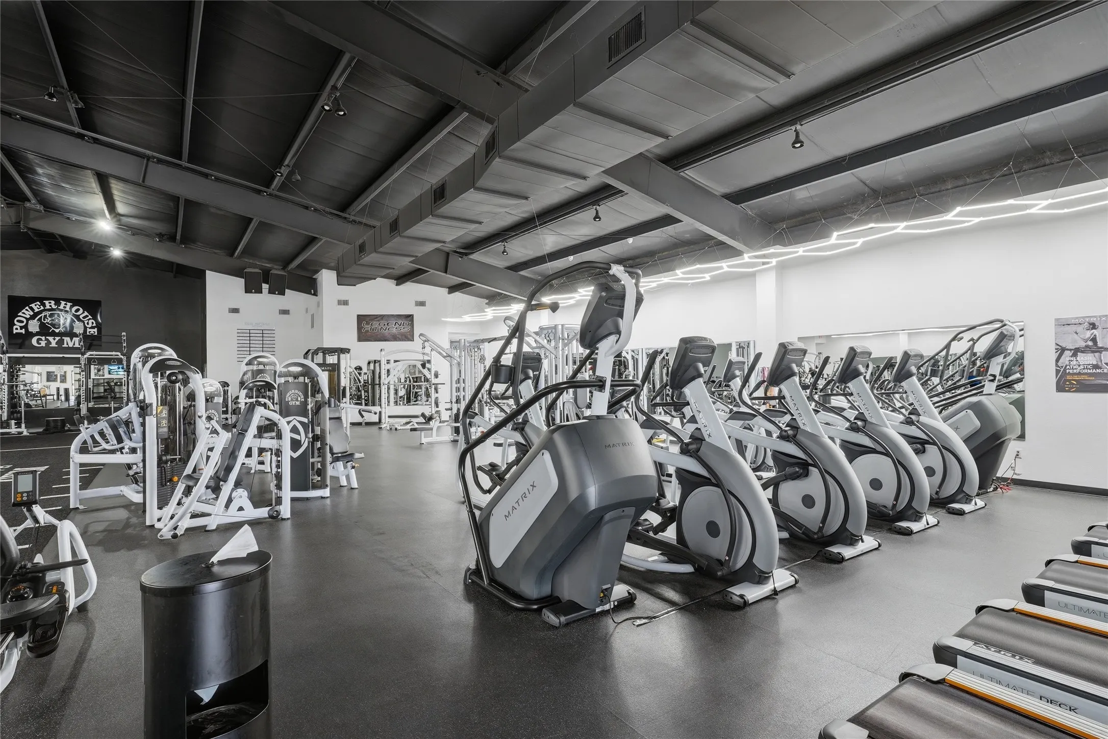 Expansive fitness area featuring multiple cardio machines, strength training equipment, a high ceiling with exposed structural beams, recessed lighting, and wall-mounted mirrors