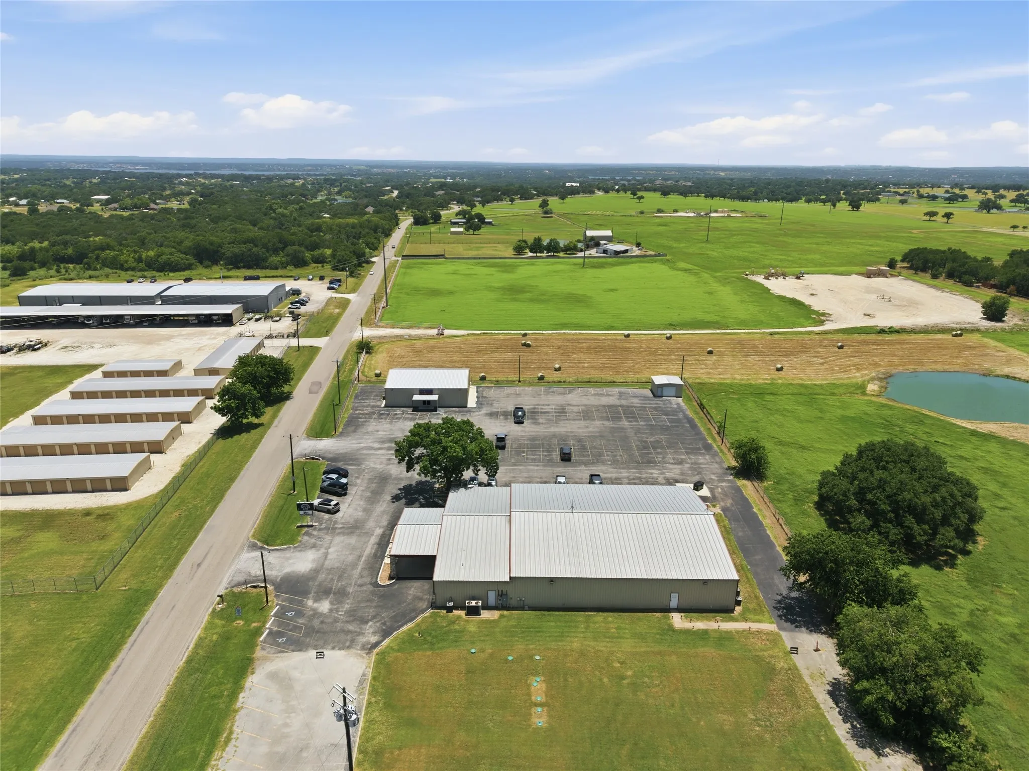 Commercial building with a metal roof and paved parking area, surrounded by expansive green fields and mature trees