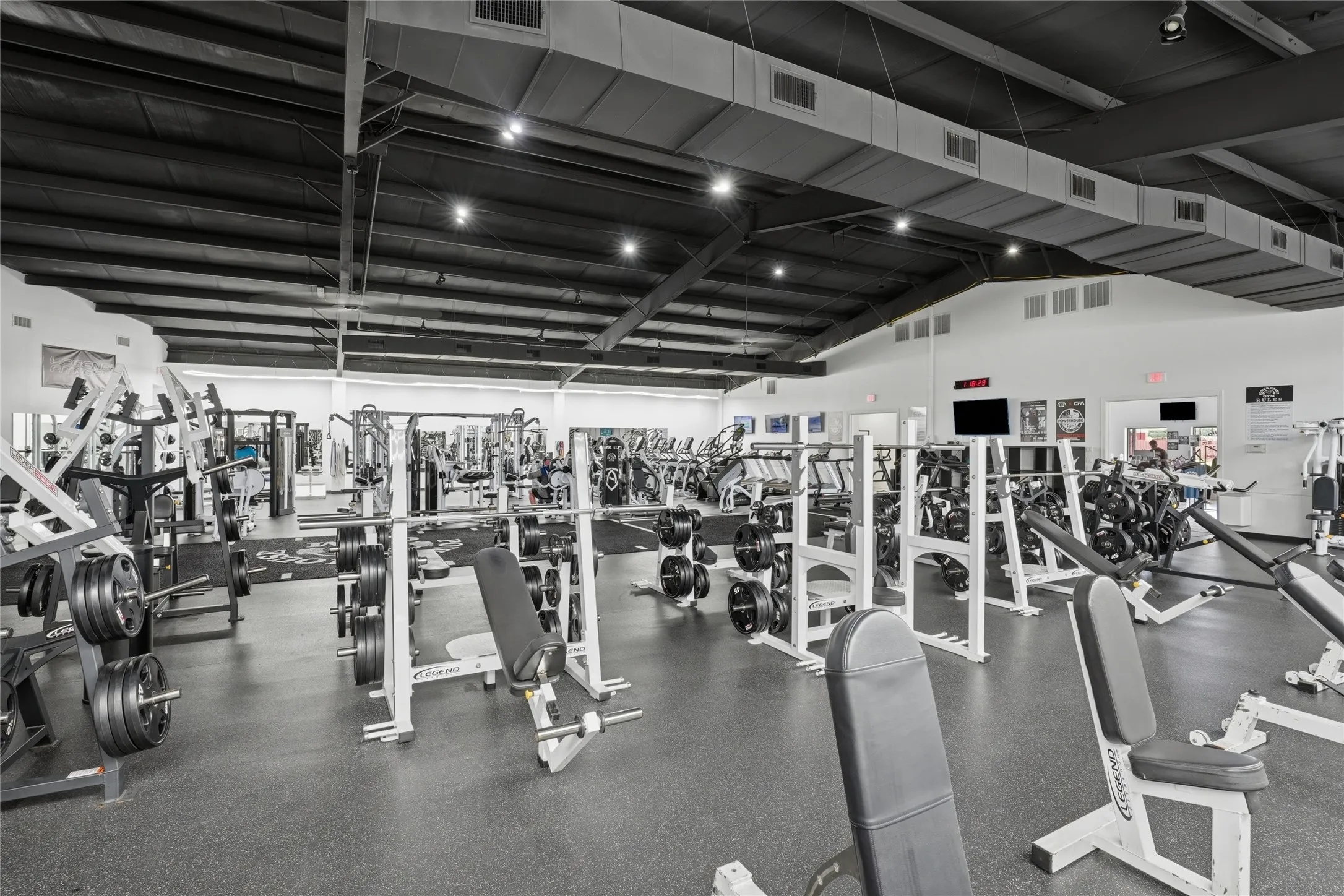 Spacious fitness area featuring a high exposed ceiling with ductwork, recessed lighting, white perimeter walls, and dark rubber flooring