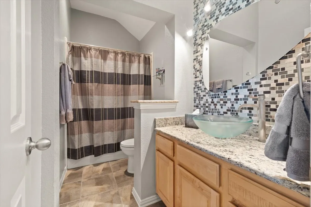 Bathroom featuring a glass vessel sink, granite countertop, mosaic tile backsplash, wood cabinetry, and a shower with tile-finish flooring