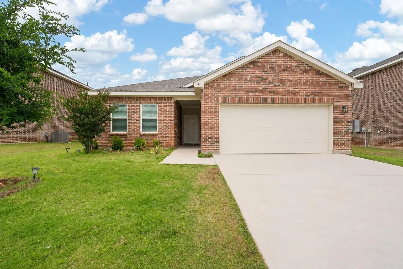 Front exterior — All-brick single-story home with covered entry, single-car garage, wide concrete driveway, and solar path lighting along the front lawn. Mature crape myrtle and established landscaping flank the entry.