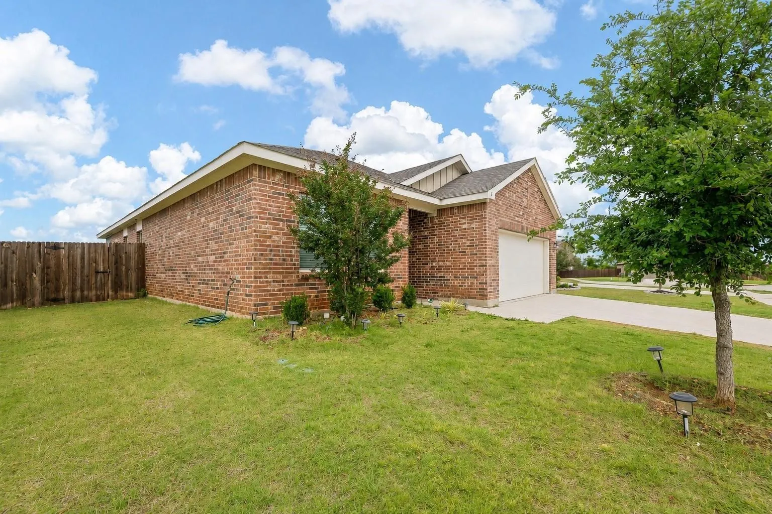 Side and rear exterior — Brick exterior with board-and-batten gable accent, single-car garage, solar path lighting, and privacy wood fence along the side yard. Established trees and low-maintenance foundation plantings throughout the side lawn.
