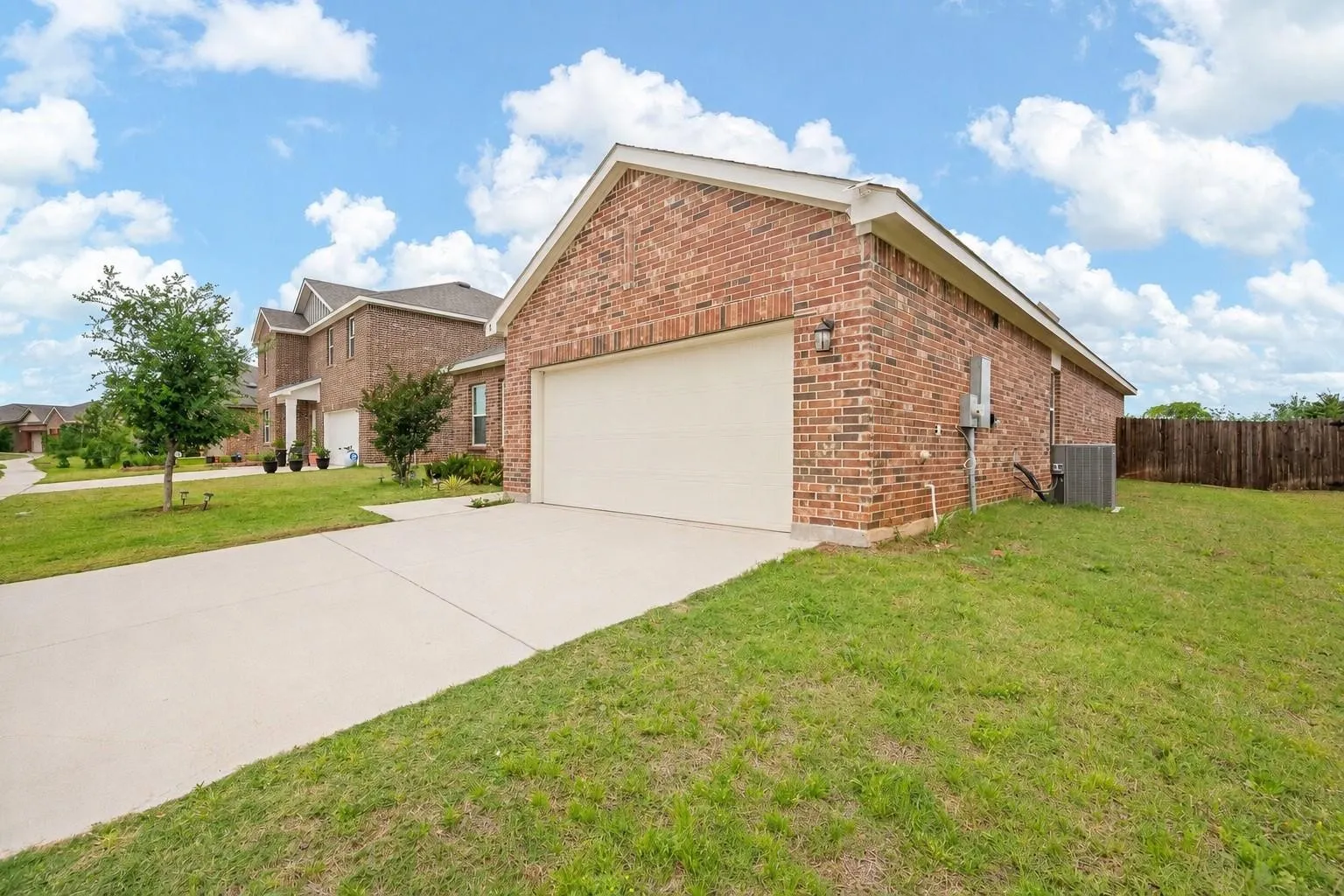 Driveway and garage approach — Wide concrete driveway with extended parking pad, single-car garage with coach light, HVAC unit on the side yard, and wood privacy fence along the rear. Established neighborhood streetscape visible in background.