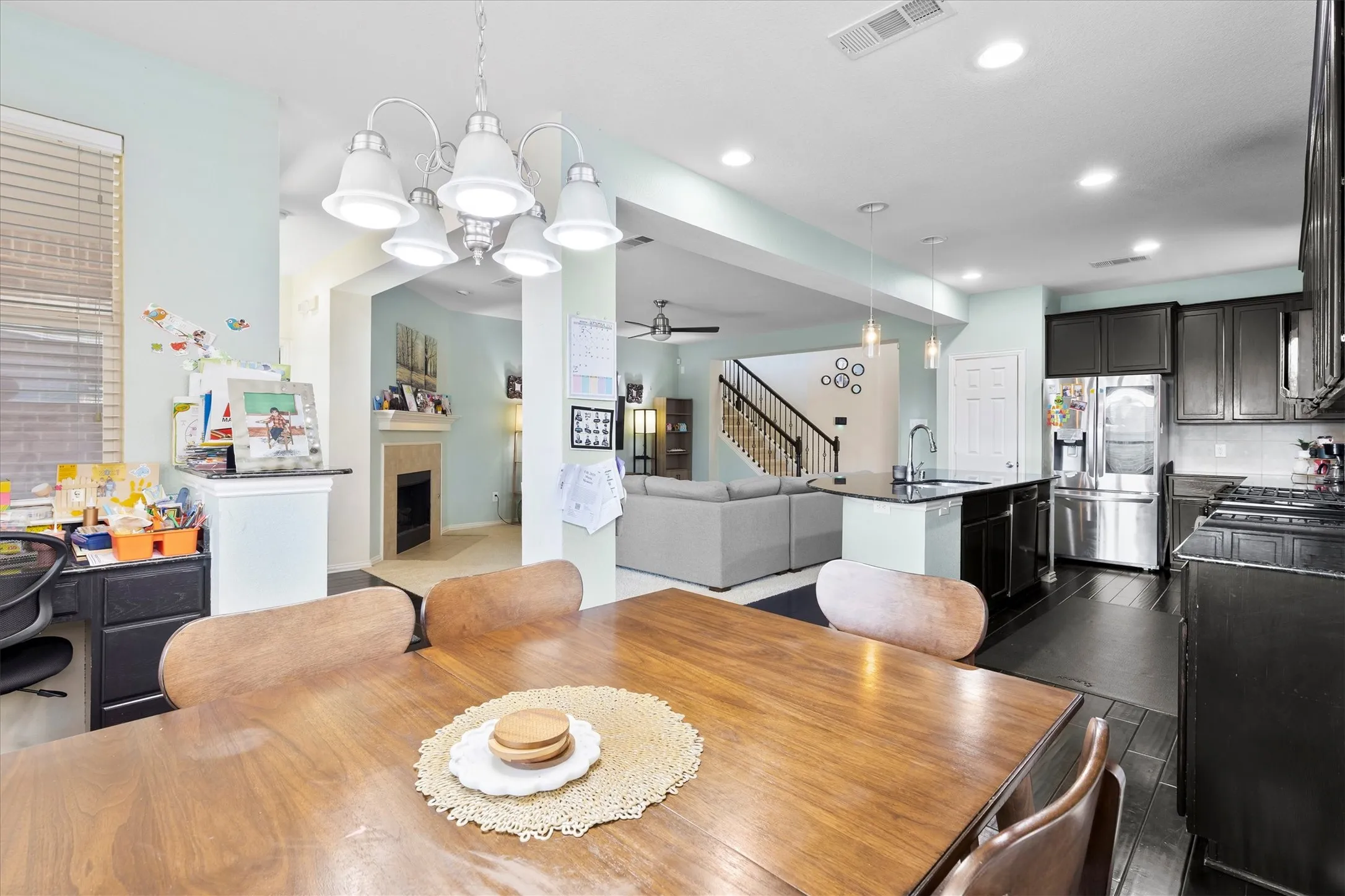 Open-concept living space featuring a dining area with a multi-light chandelier, a kitchen with dark cabinetry and stainless steel appliances, a center island with a sink, and a living area with a fireplace visible in the background