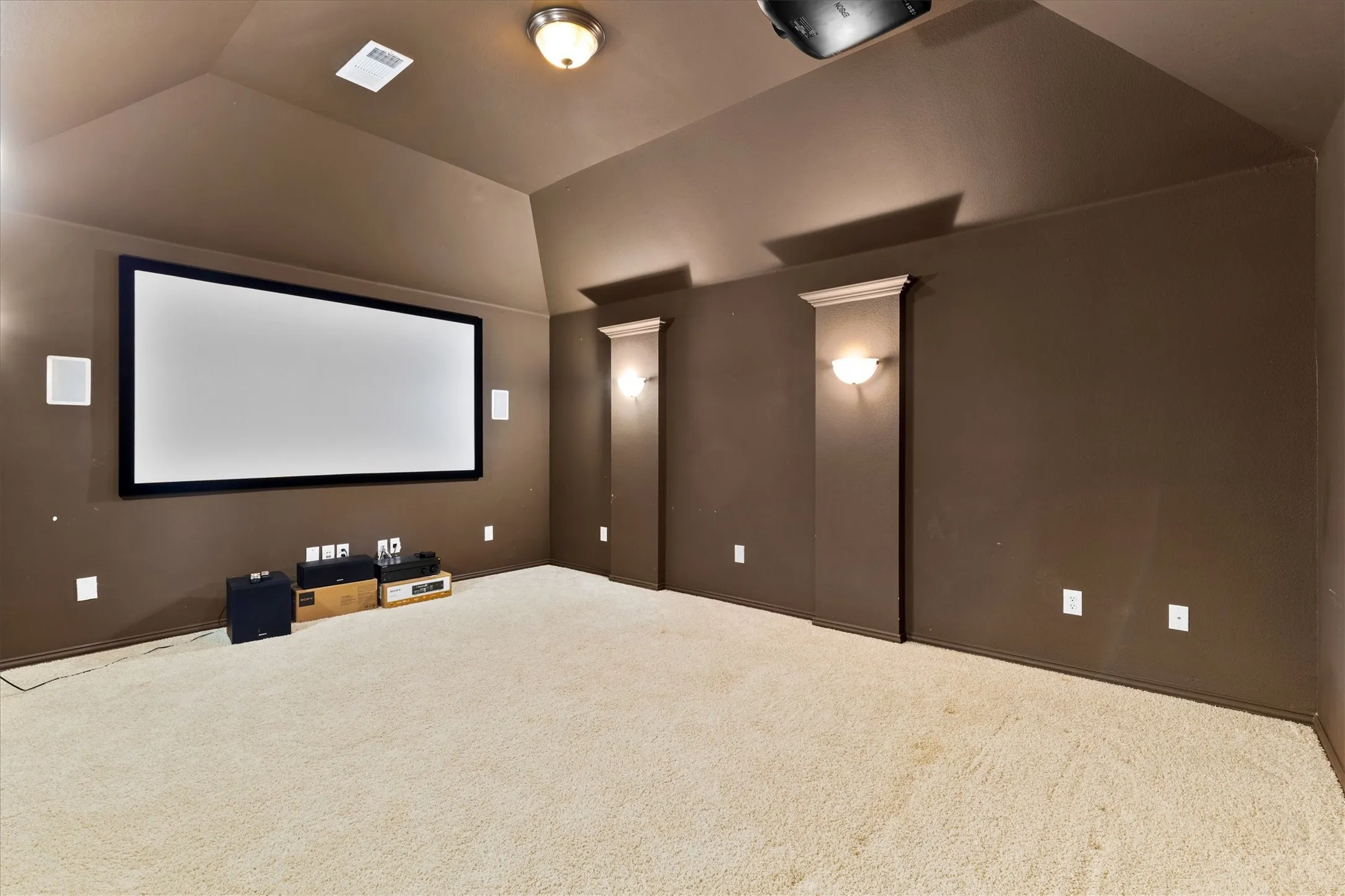 Dedicated media room featuring a large projection screen, recessed ceiling projector, wall-mounted speakers, and light-colored carpet flooring