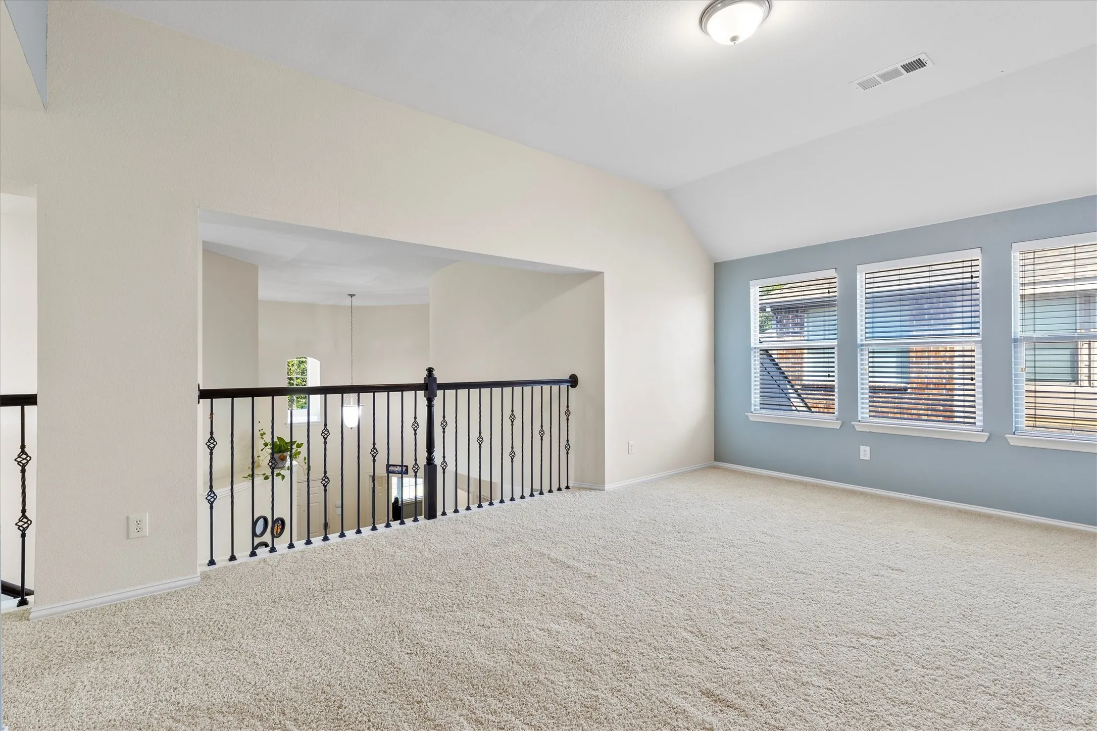 Carpeted loft space featuring an open railing overlooking the area below, three windows with horizontal blinds, a vaulted ceiling, and a flush mount light fixture