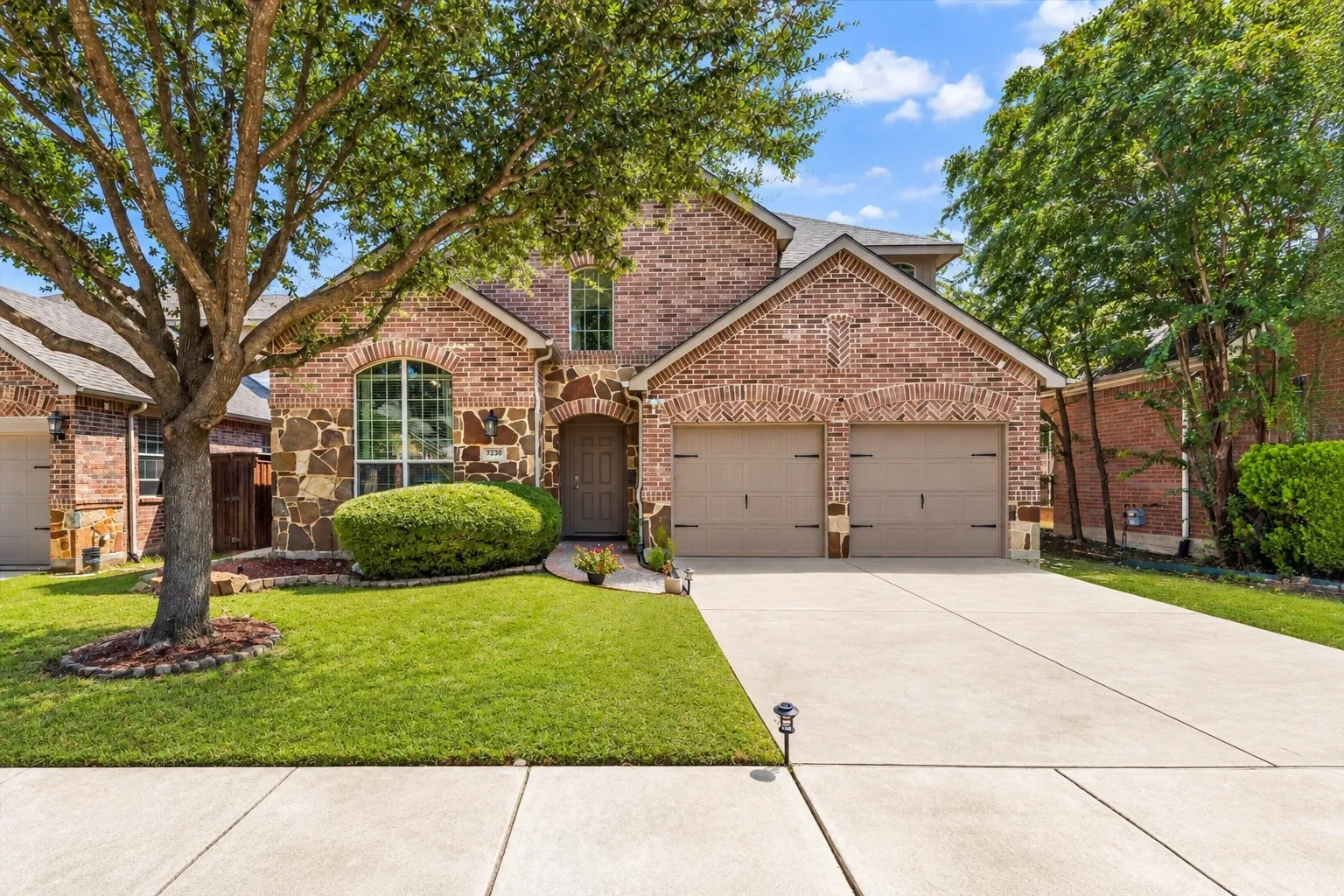 Brick and stone facade with an arched entry, mature tree, manicured lawn, and a two-car garage