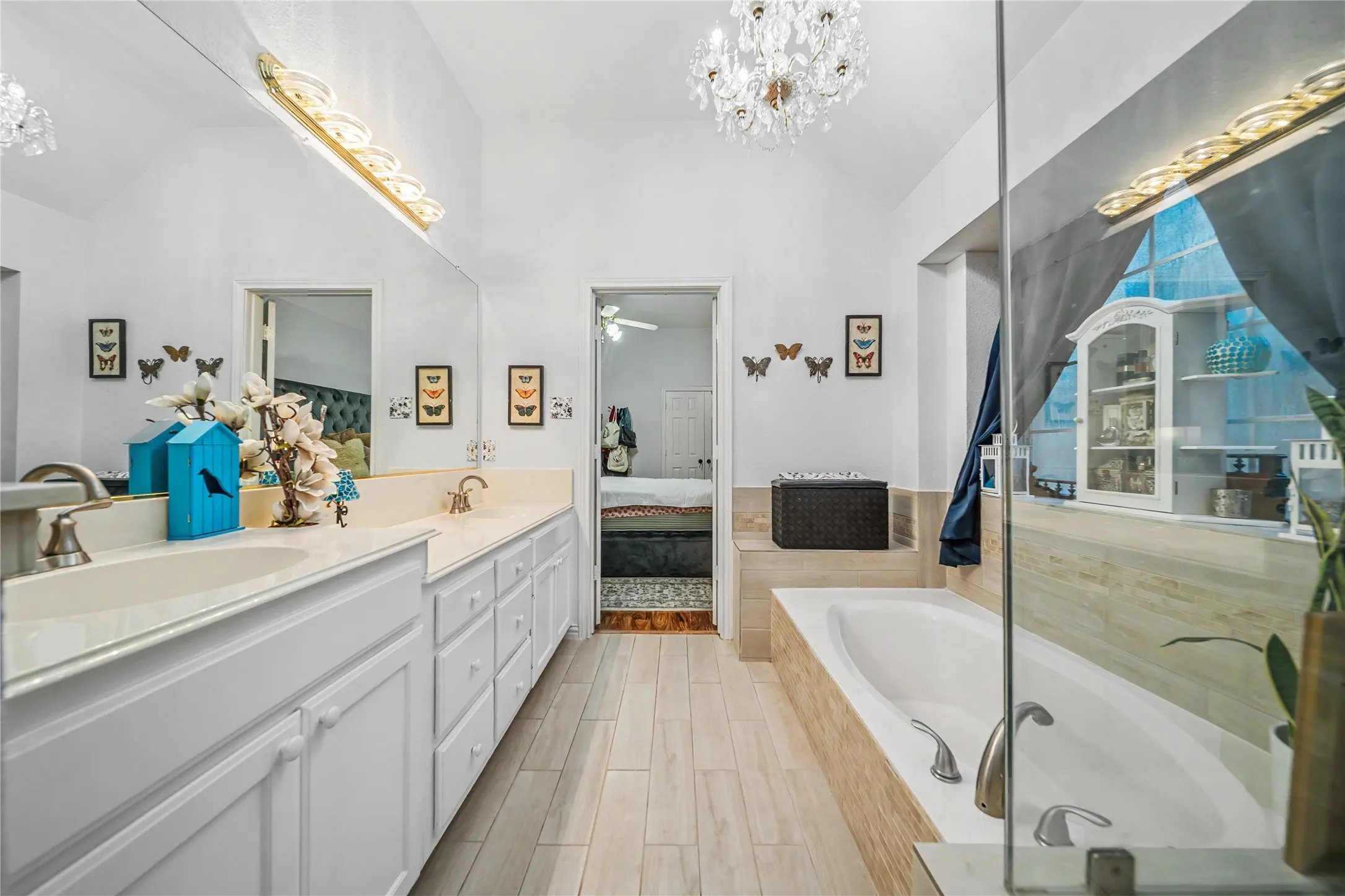 Spacious bathroom featuring a dual vanity with white cabinetry and light-toned countertops, wood-finish flooring, a built-in bathtub with tiled surround, and a crystal chandelier