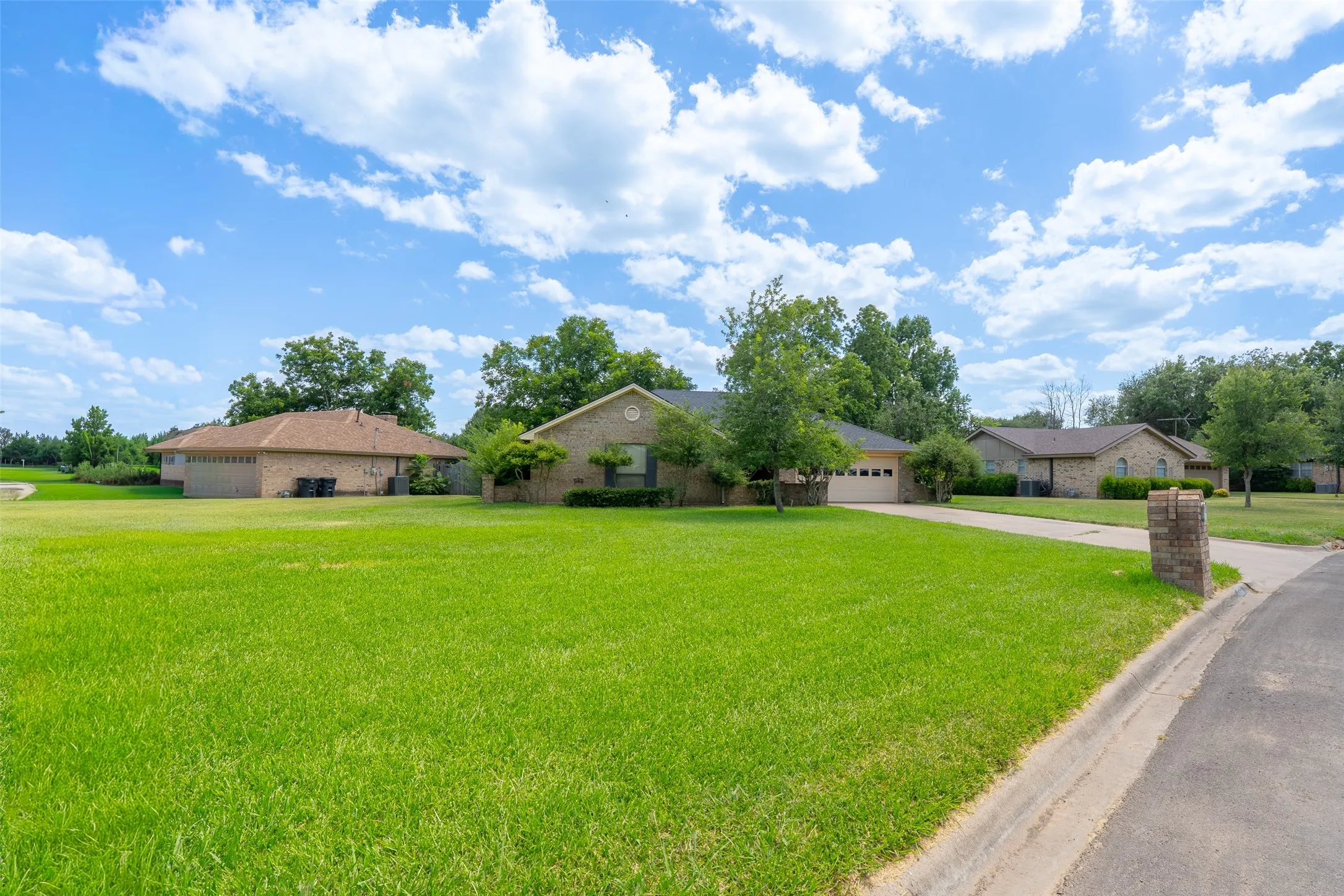 Expansive green lawn with mature trees and a paved driveway, featuring a brick-clad residence with an attached garage