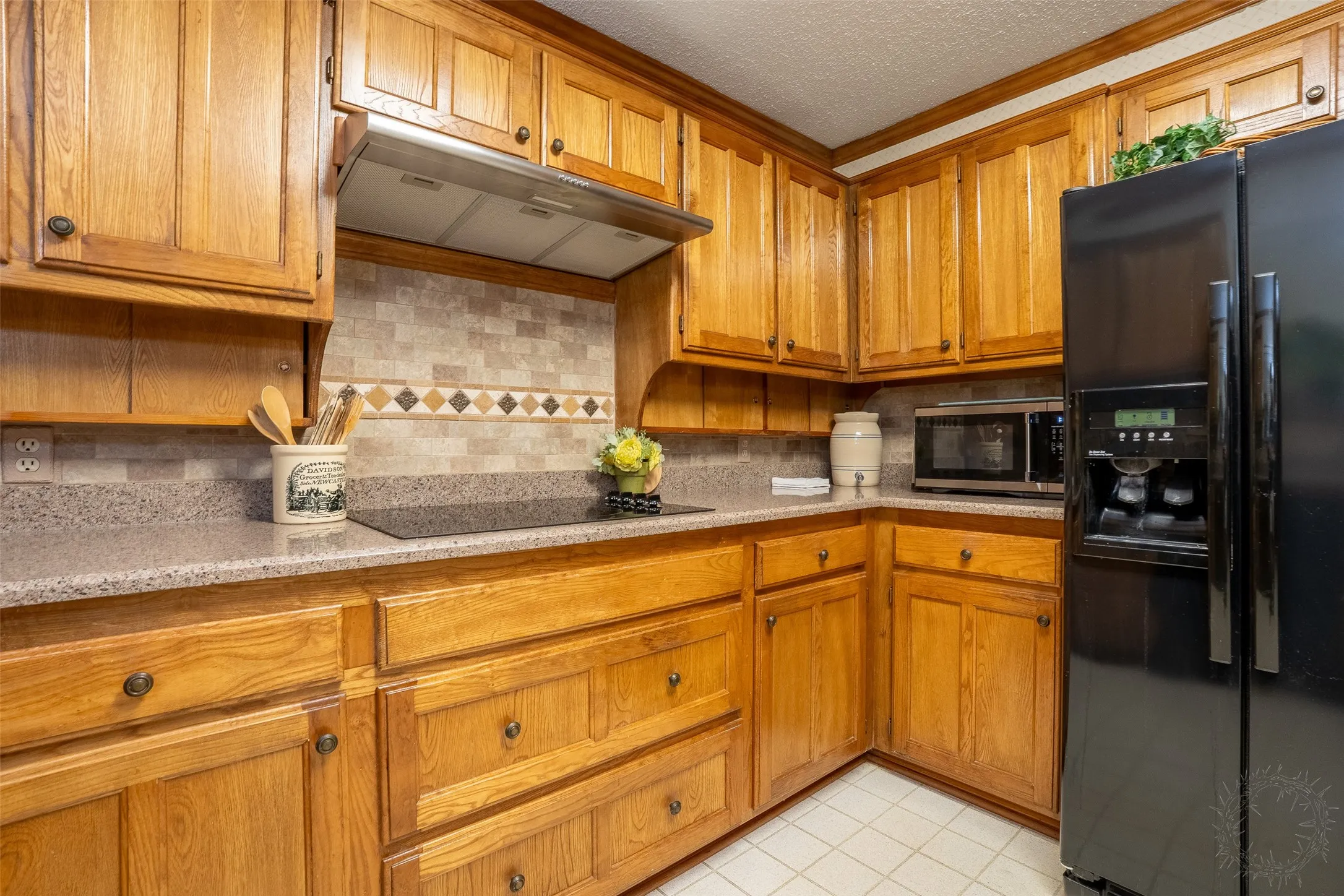 Kitchen featuring wood cabinetry, stone-look countertops, tiled backsplash with decorative accents, and a built-in cooktop
