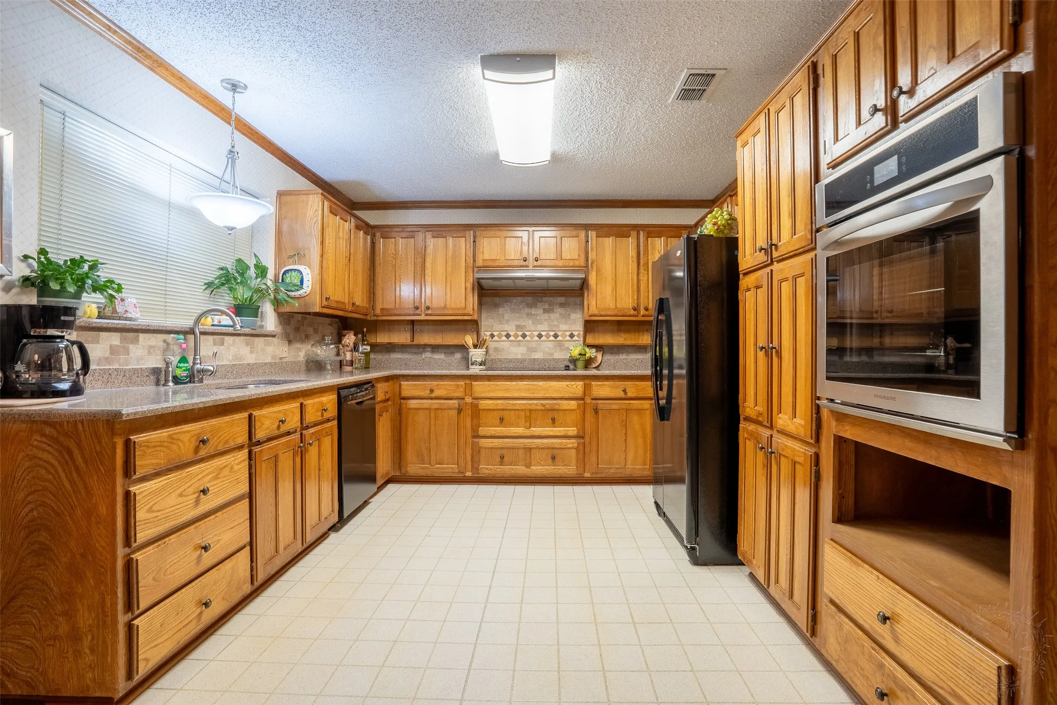 Kitchen featuring extensive wood cabinetry, light-toned tiled flooring, built-in stainless steel double ovens, a stainless steel sink, and a patterned tile backsplash