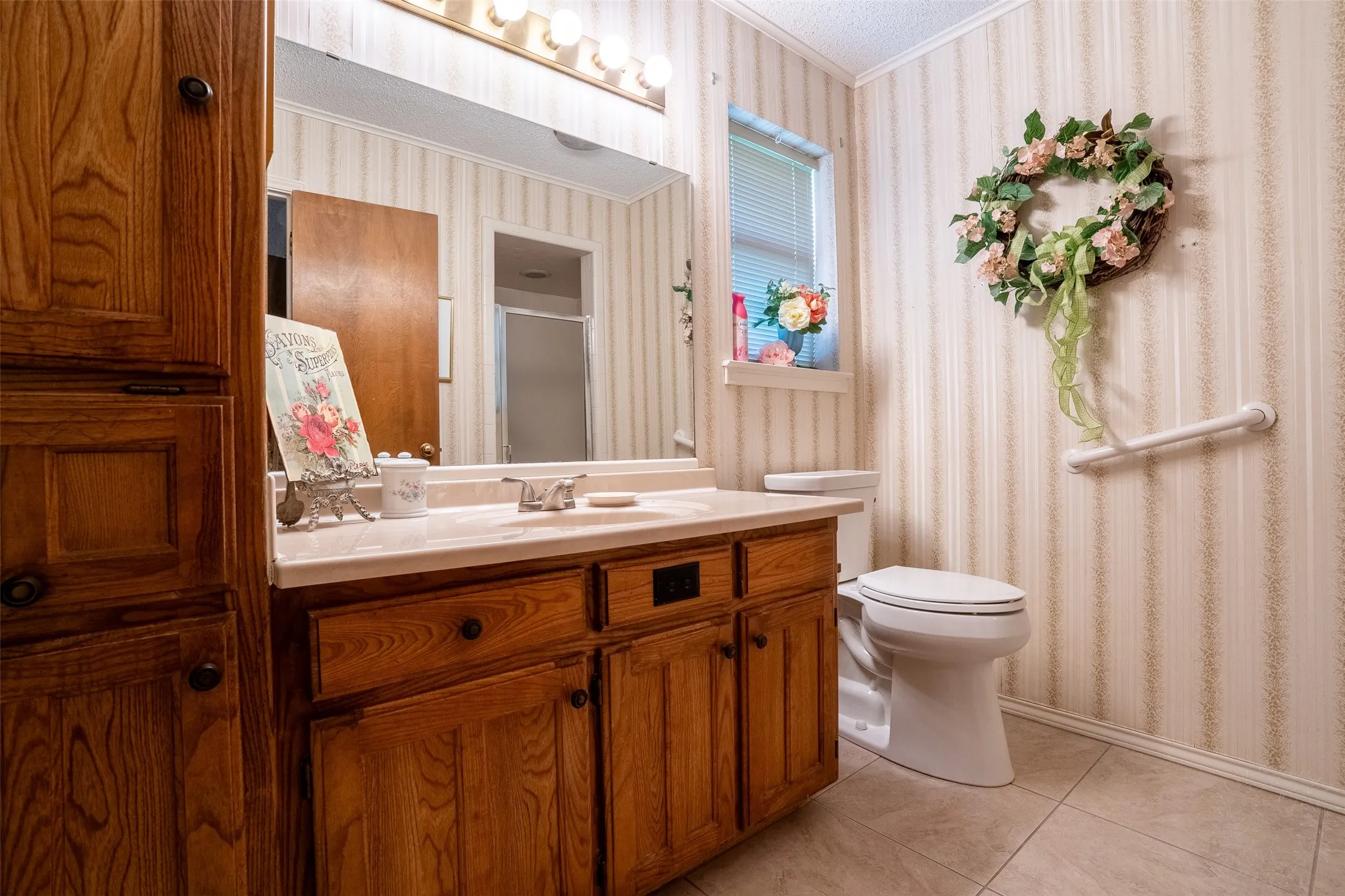 Bathroom featuring a wood-finish vanity with a light-toned countertop, a built-in wood-finish linen cabinet, striped wallpaper, and tiled flooring