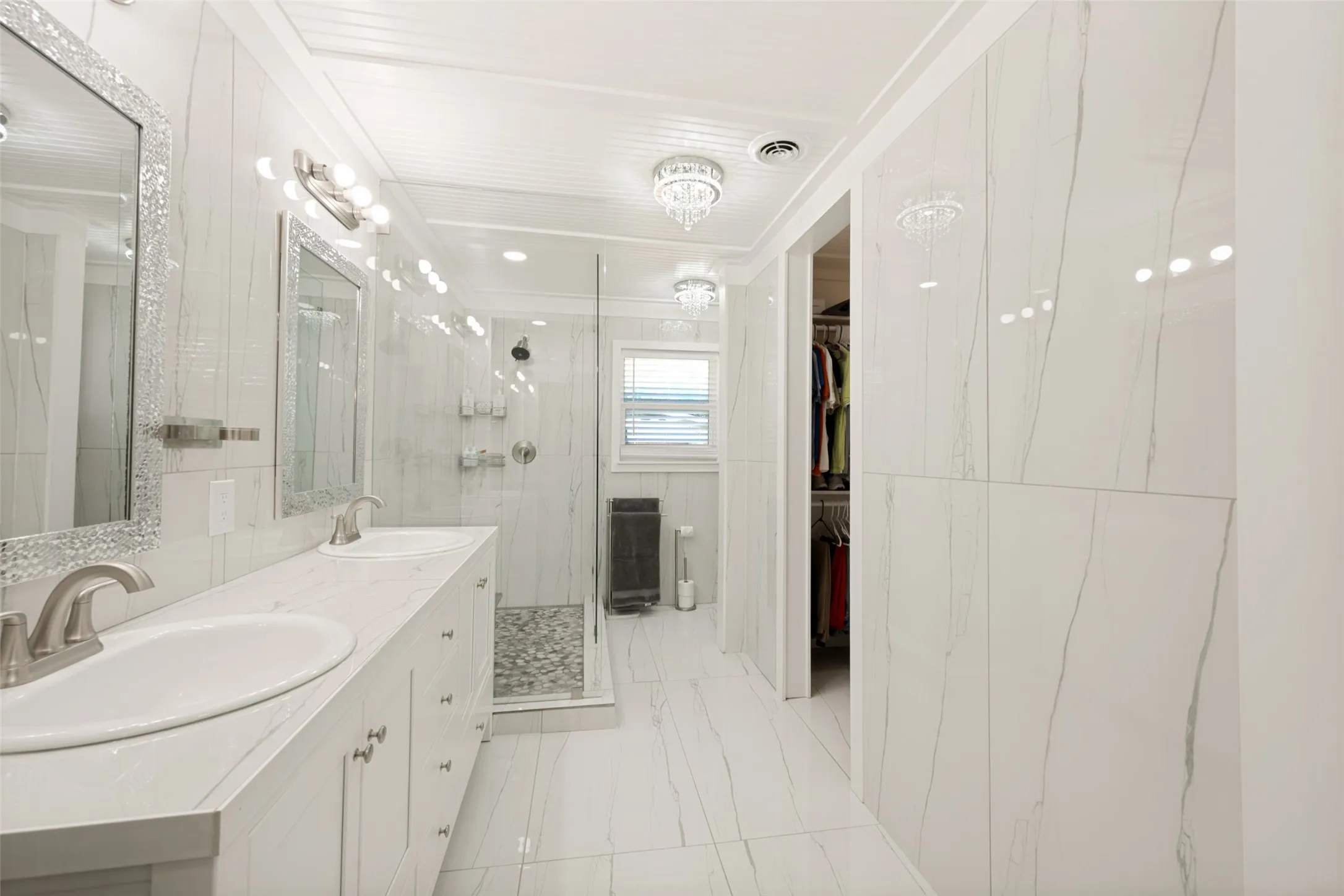 Spacious bathroom featuring a dual vanity with undermount sinks and brushed nickel faucets, a glass-enclosed shower with pebble flooring, white large-format wall and floor tiles, and a chandelier-style light fixture