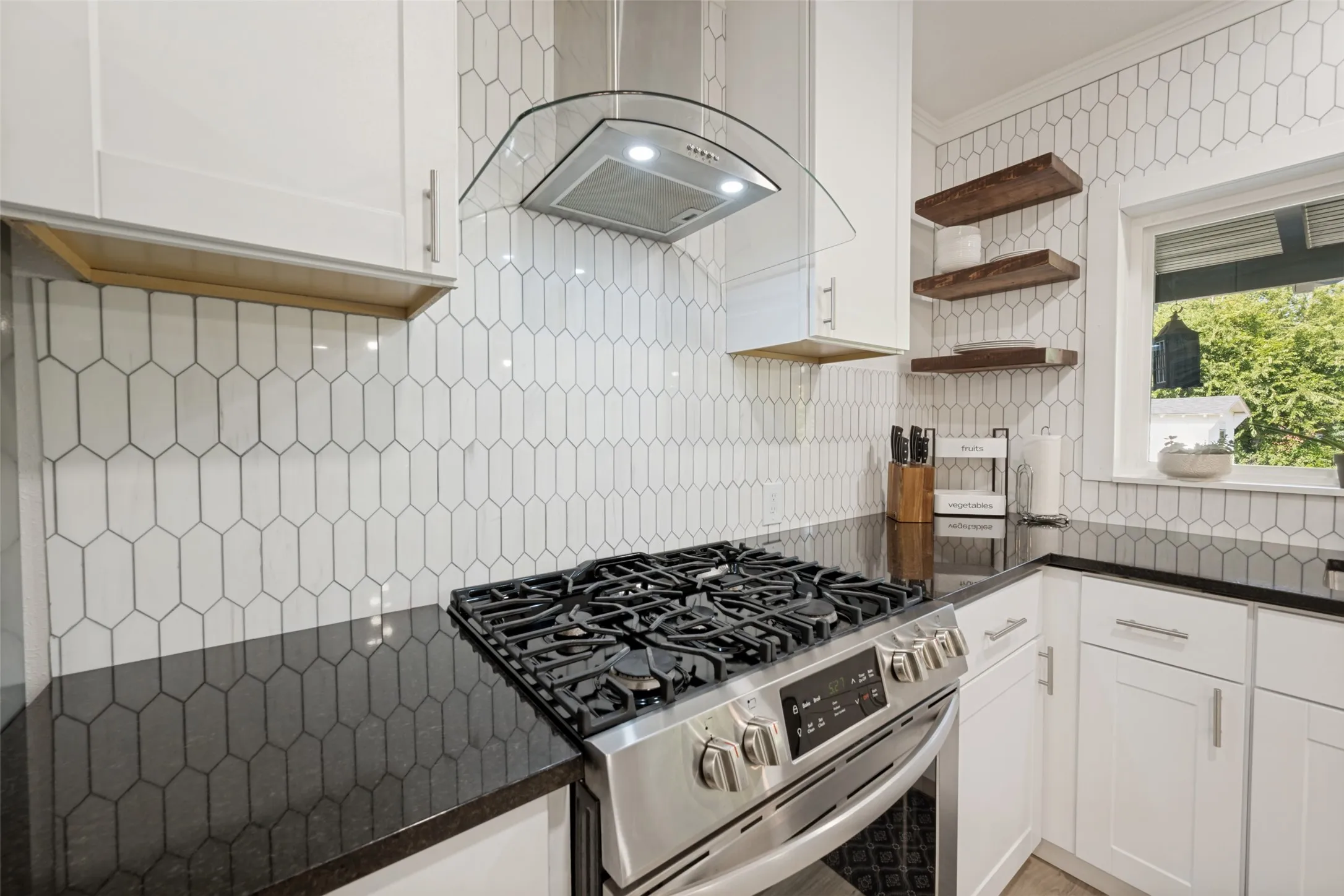 Stainless steel range with a gas cooktop, black countertops, custom white cabinetry, glass and stainless steel range hood, and a geometric tile backsplash