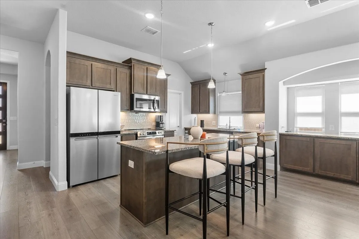 Kitchen featuring a granite-topped island with pendant lighting, stainless steel appliances, wood-finish cabinetry, a tiled backsplash, and wood-finish flooring