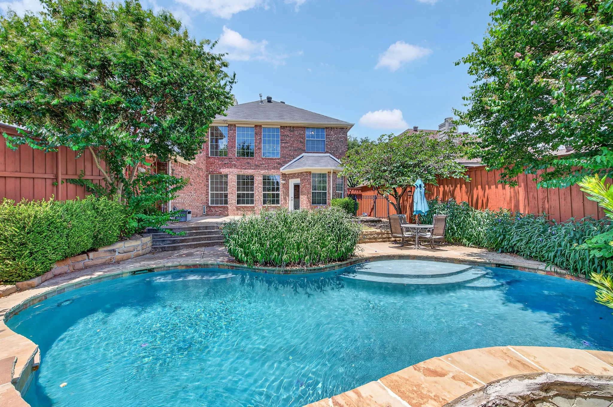 Private backyard featuring a large swimming pool with stone coping and integrated steps, a brick-facade residence, mature trees, and a wooden perimeter fence